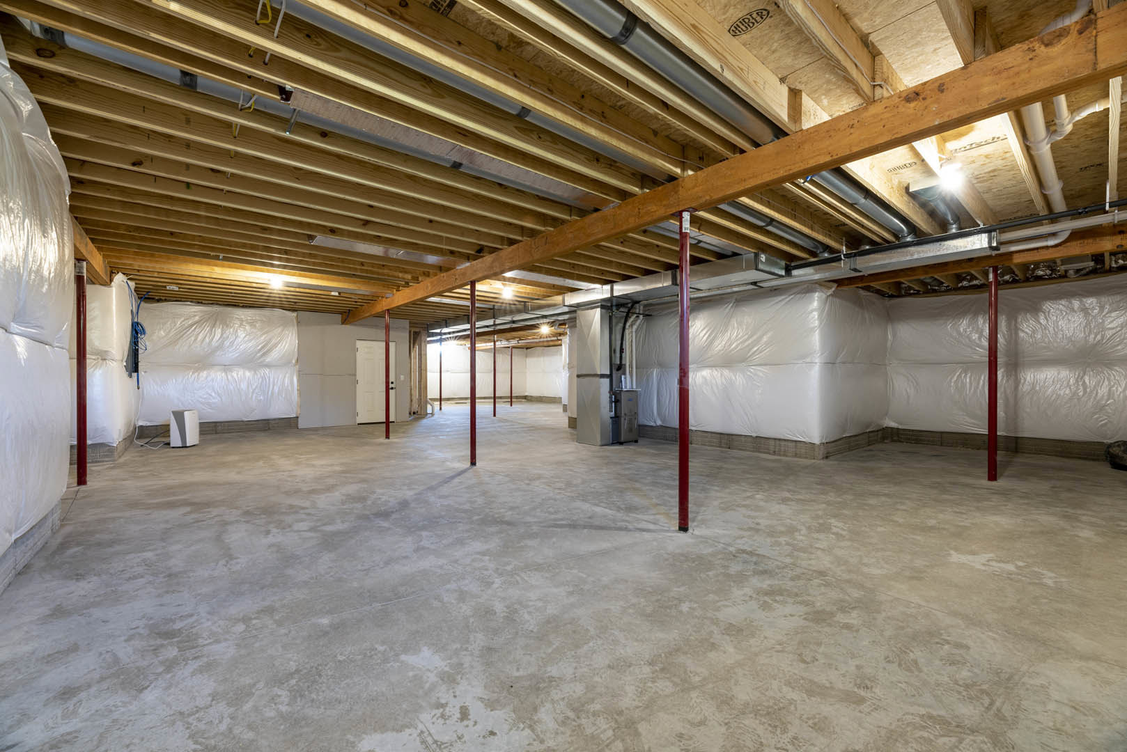 White-walled room with exposed wooden ceiling beams, concrete floor with red support poles, visible pipes overhead, white rectangular object on wooden surface, logo on a nearby