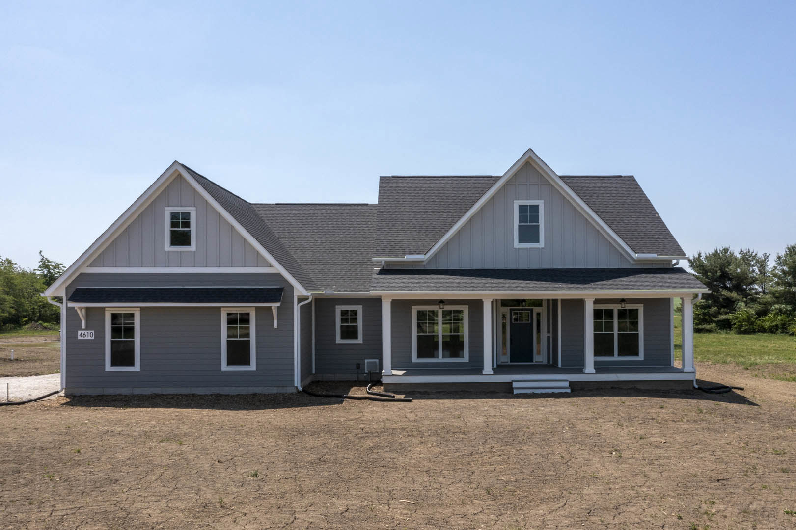Two-story house with grey roof, white-framed square windows, light siding, covered porch, and dirt yard in foreground