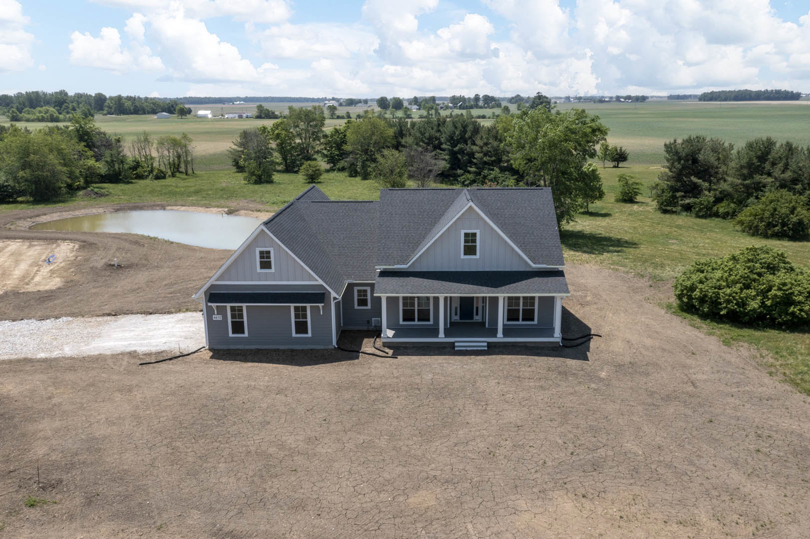 Two-story house with light siding and gabled roof, manicured lawn in front, large bush to the side, pond and grassy field with scattered trees in the background under a partly
