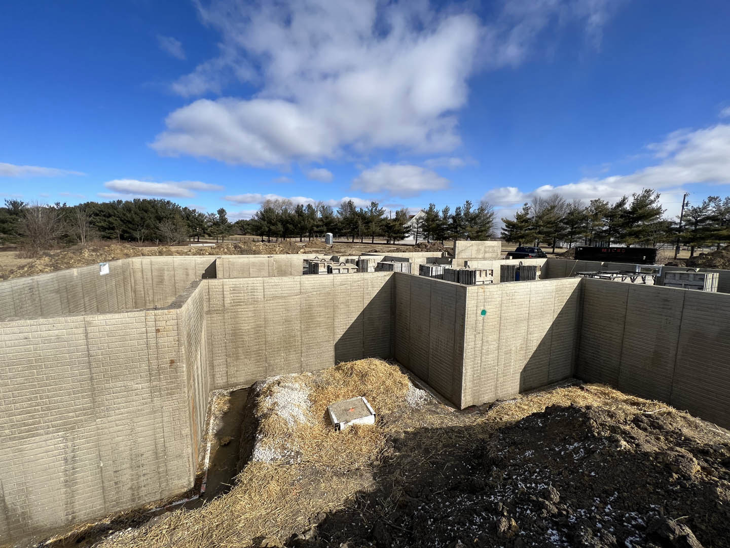 Concrete block walls and foundation under construction, surrounded by piles of dirt and hay, leafless bush, grassy ground, and trees in the background beneath a partly cloudy blue