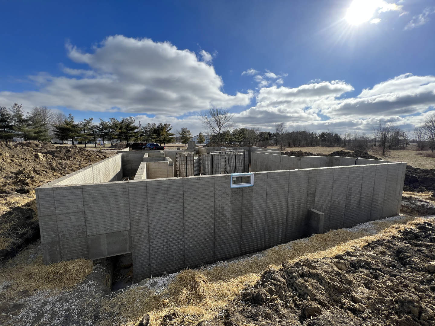 Concrete exterior wall with rectangular window, unfinished construction, blue sky with scattered clouds, leafless tree nearby, pile of hay beside brick wall