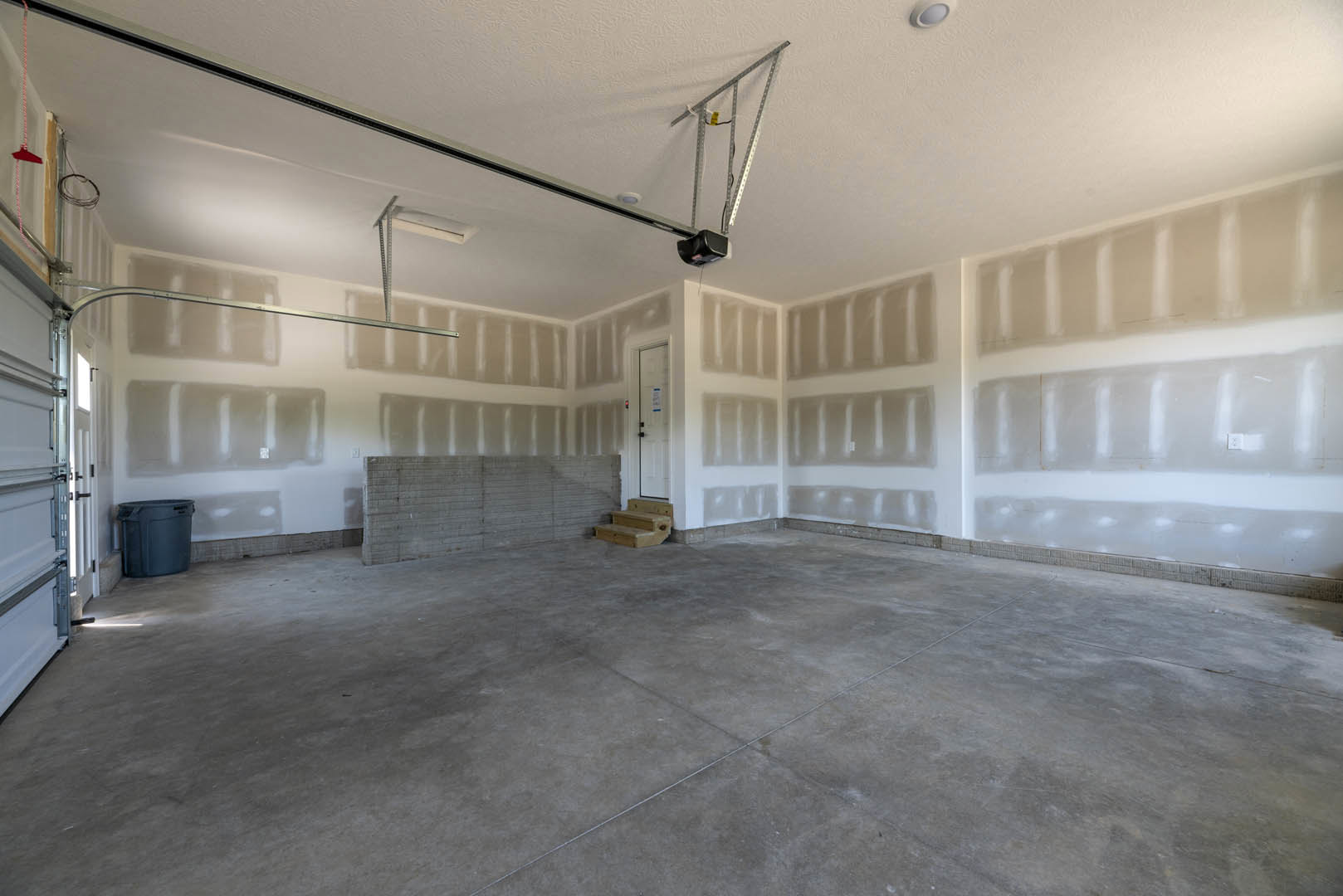 Concrete floor with a white door, exposed brick wall, wooden staircase, and a grey plastic bin in an empty room