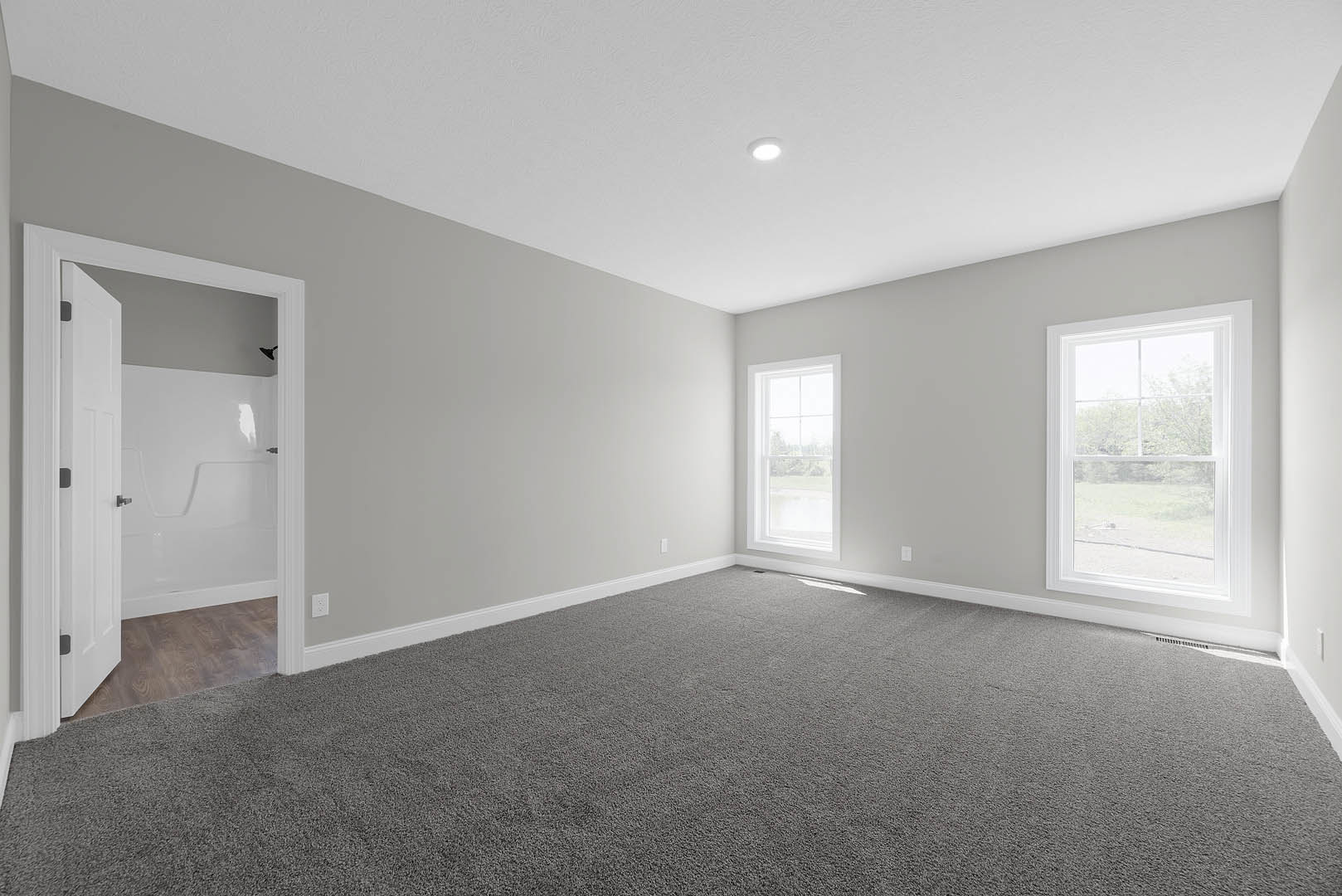 Room with grey carpet, open white door, large windows showing outdoor greenery, white walls, and ceiling with recessed lighting