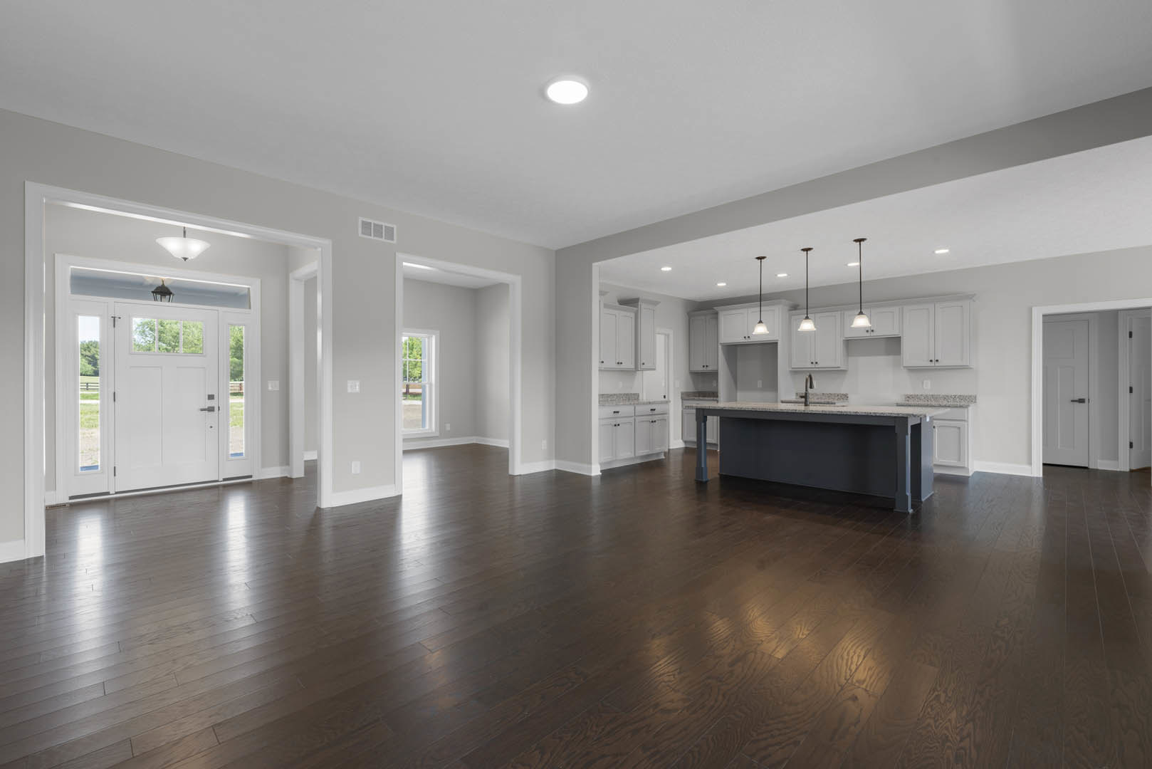 Spacious open floor plan featuring a central kitchen island with sink, hardwood flooring throughout, white cabinetry, ceiling lights, and a white door with black handle and window