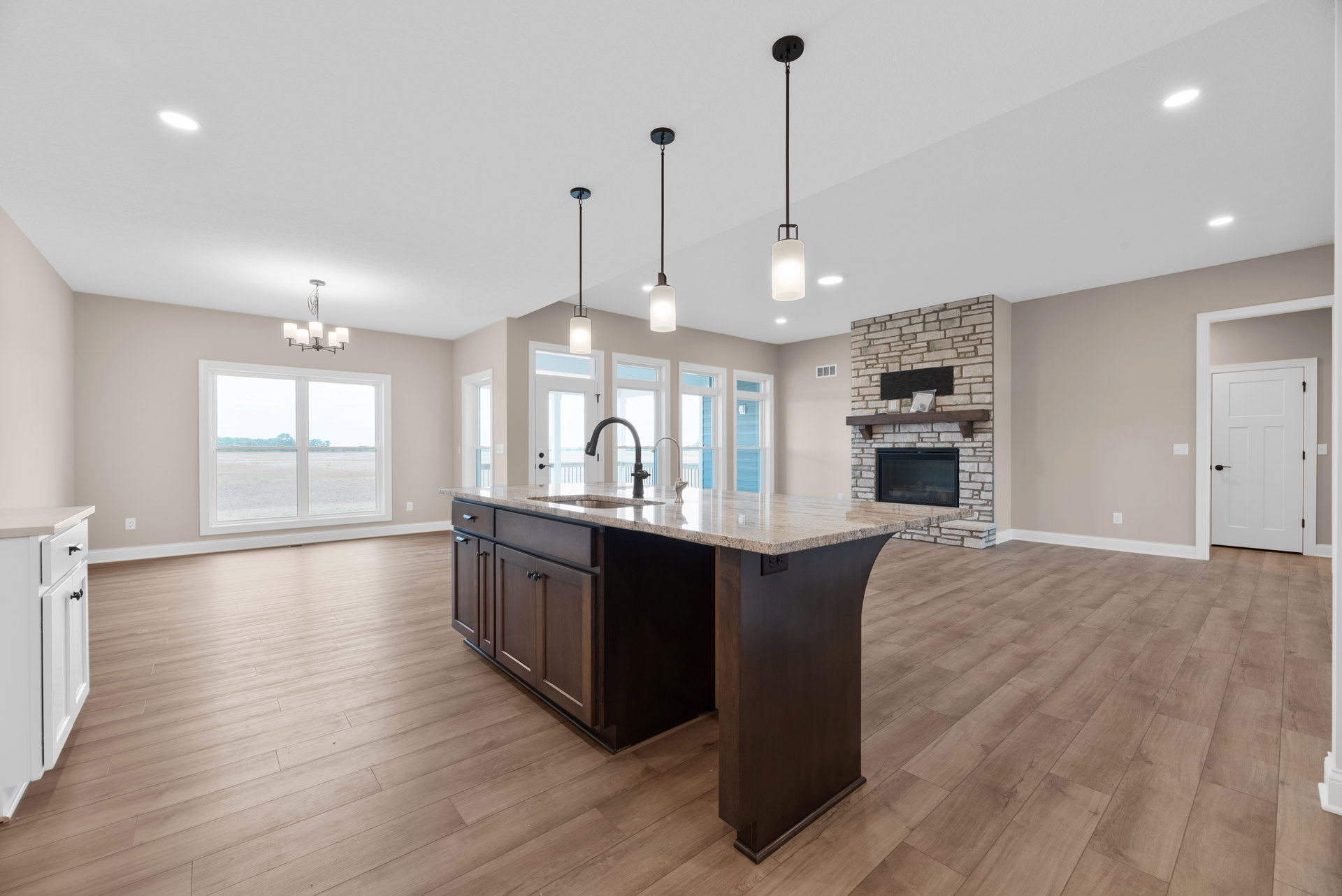 Marble-topped kitchen island with built-in sink, surrounded by white cabinetry and laminate flooring, large window overlooking a field, white door with black handle, recessed