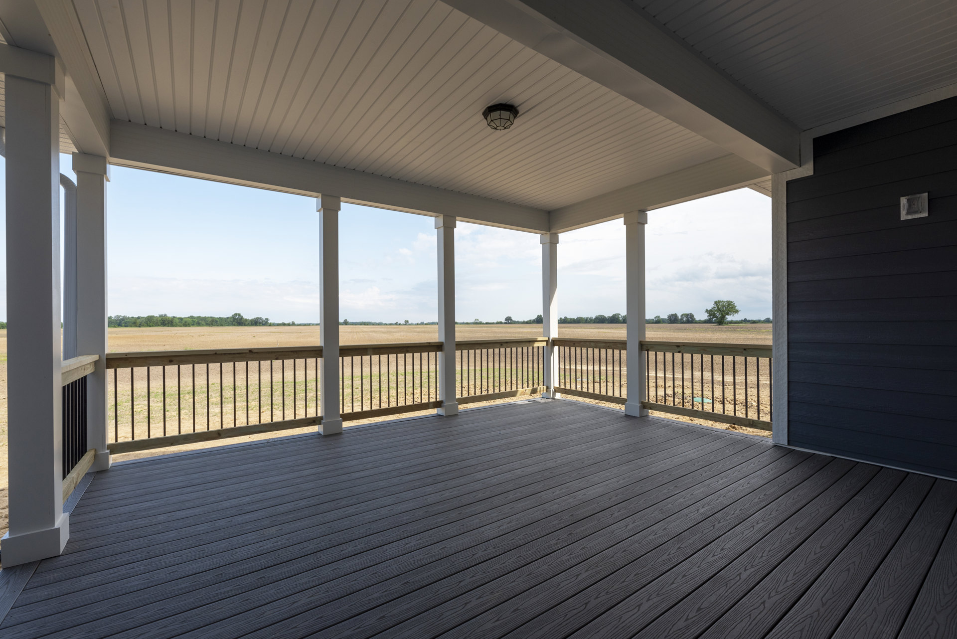 Spacious composite deck with metal railings overlooking open grassy field, rectangular pillar, ceiling light fixture, and electrical socket visible