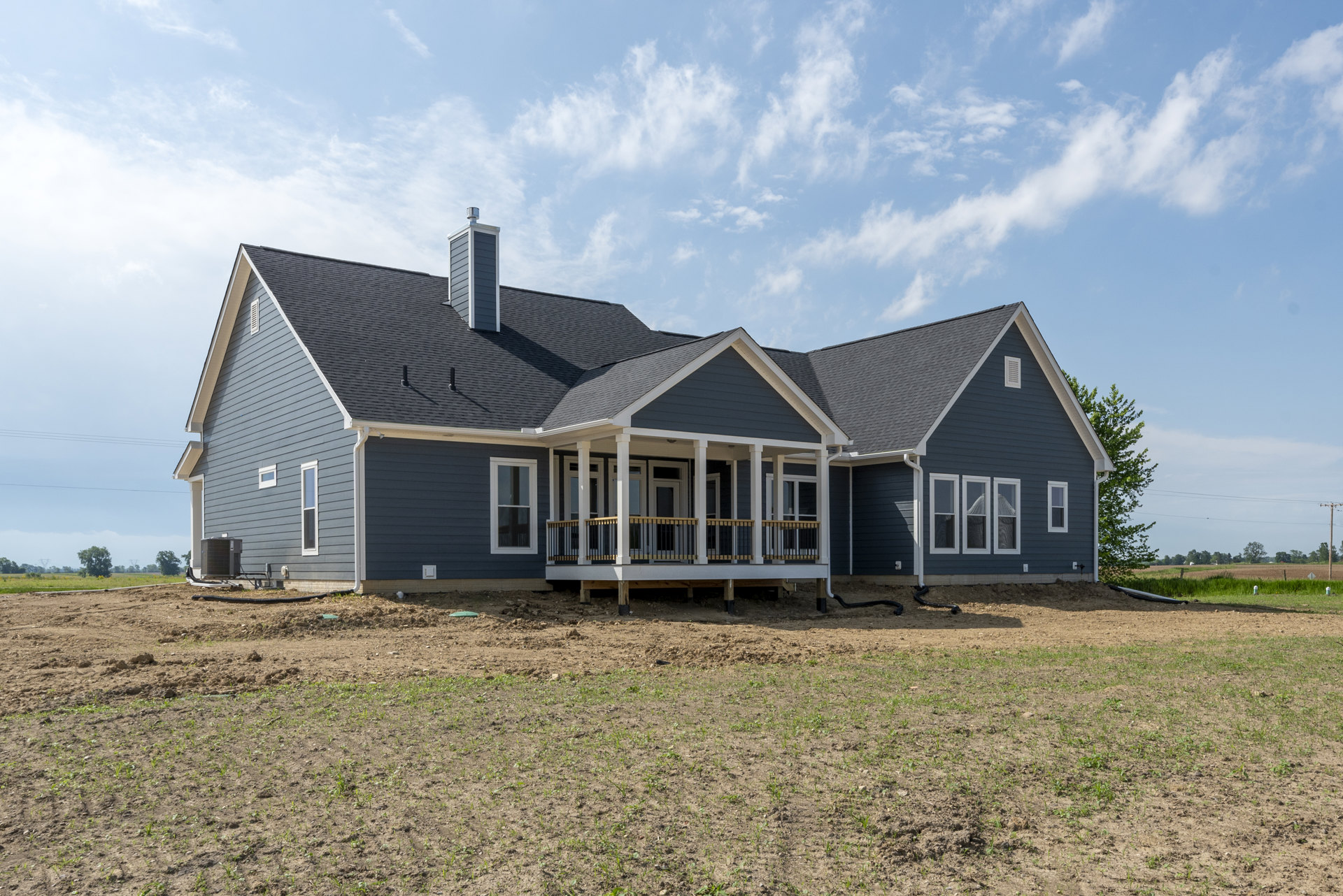 Two-story house under construction with white-framed windows, covered porch, brick chimney, and large grassy yard; black heat pump unit beside exterior wall
