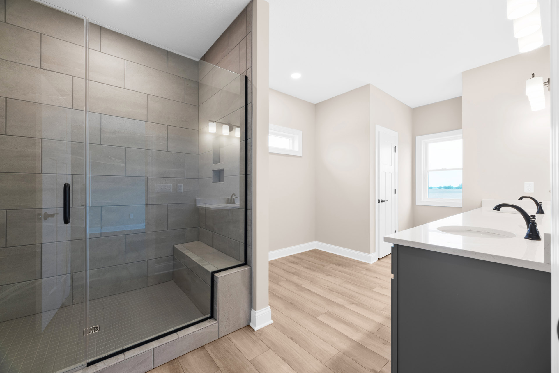 Bathroom featuring a frameless glass shower enclosure, white ceramic sink with chrome faucet, large window overlooking water, light gray tile flooring, and neutral wall tiles