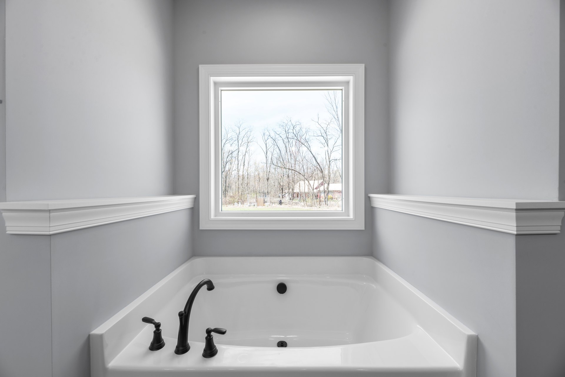 White freestanding bathtub with matte black faucet beneath large window overlooking trees, white walls and ceiling with recessed light visible