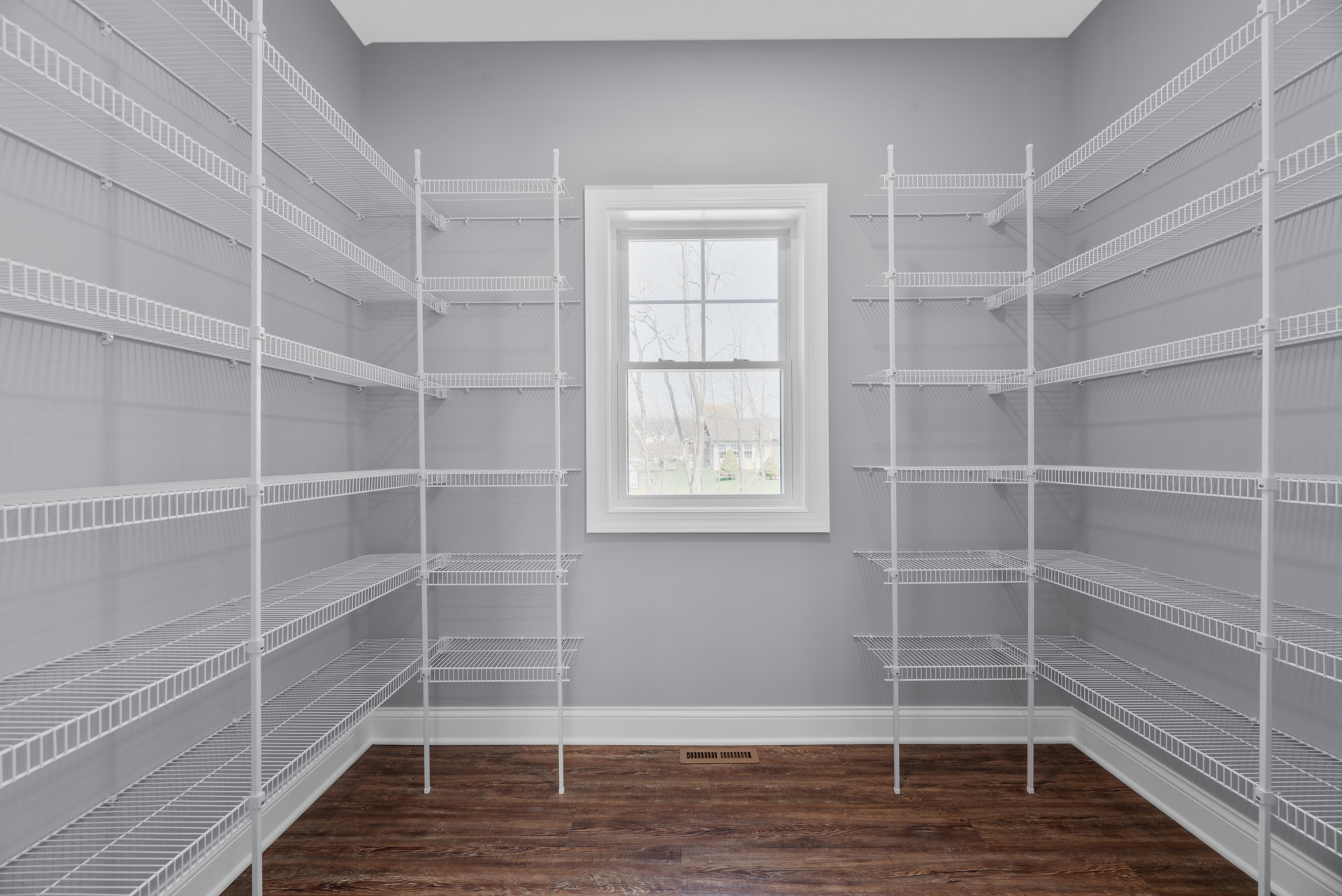 White wire shelving unit along plaster wall, wood floor with vent, large window overlooking trees