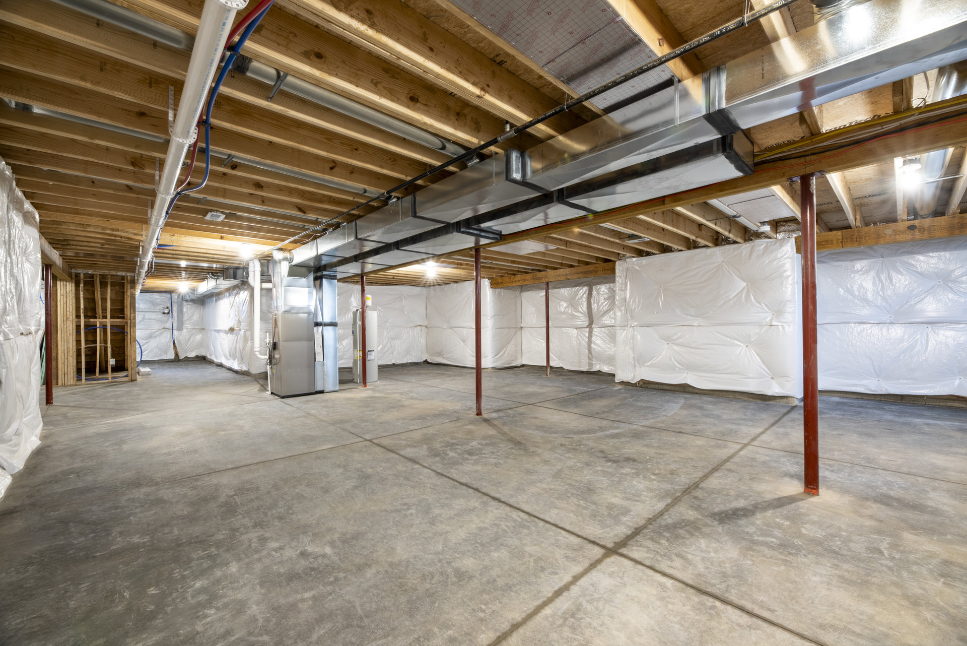 Concrete basement with exposed metal pipes, metal utility box, white plastic sheeting covering walls and floor, white plastic bag tied to vertical pole, unfinished ceiling beams