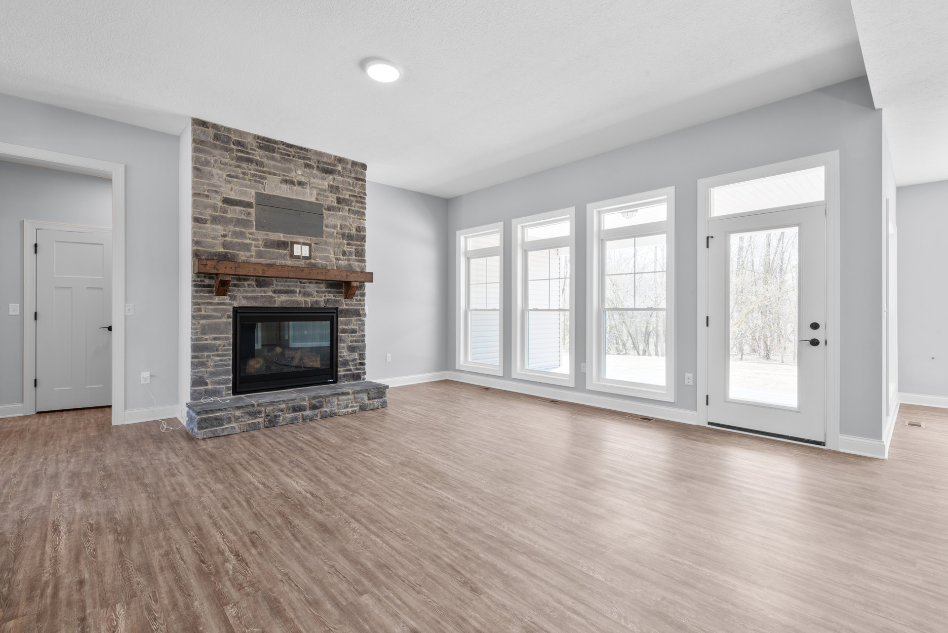 Living room with hardwood floors, glass-door fireplace, large windows showing trees, and white door with black handle