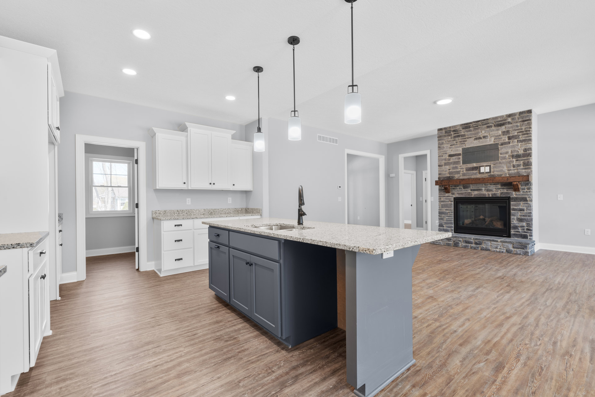 Open kitchen featuring a stone fireplace with glass doors, large central island with white countertop, wood cabinetry, stainless steel sink, and windows overlooking trees.