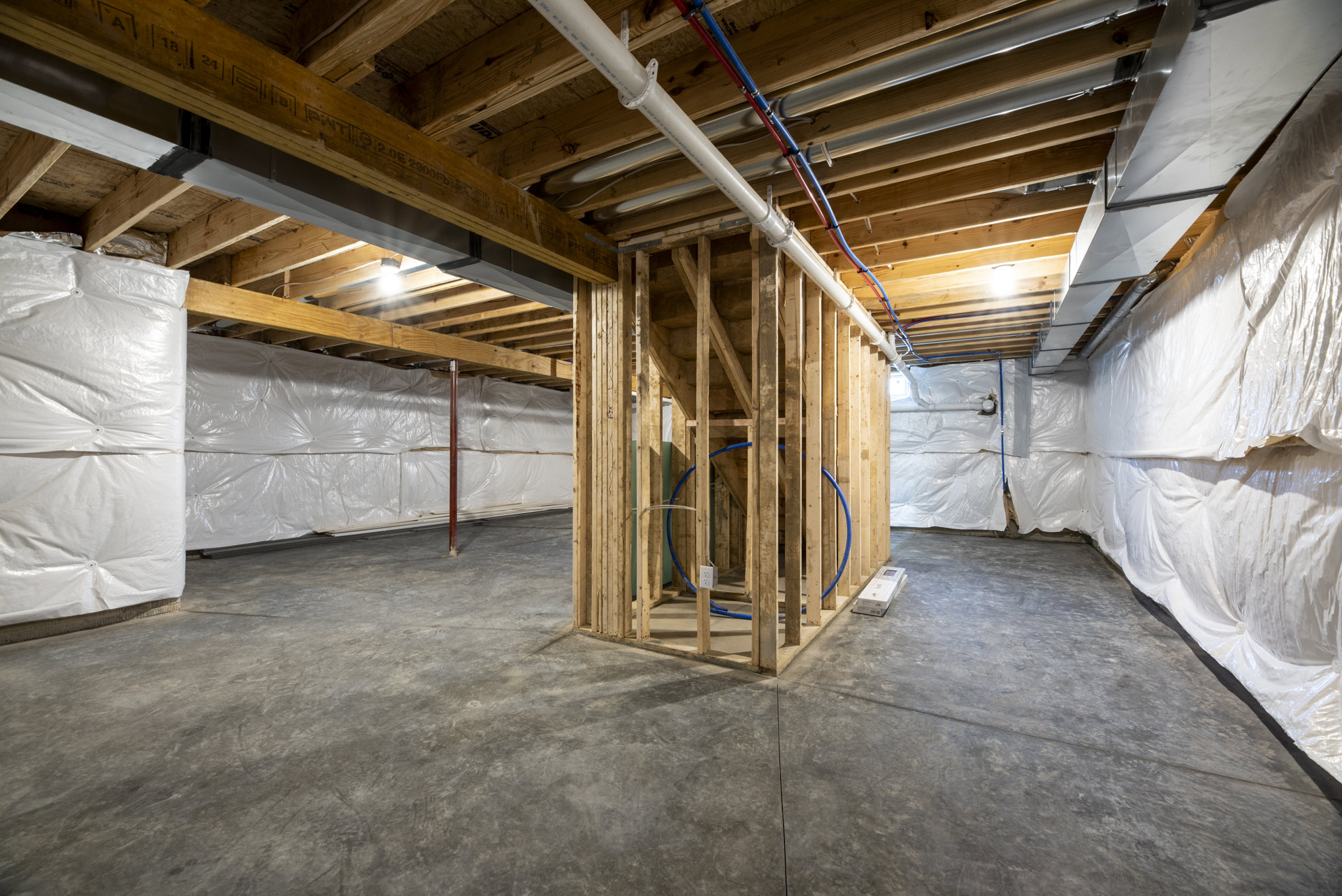Basement room with exposed wood framing, visible pipes, white plastic sheet with cutout, white box on concrete floor, unfinished walls and ceiling.