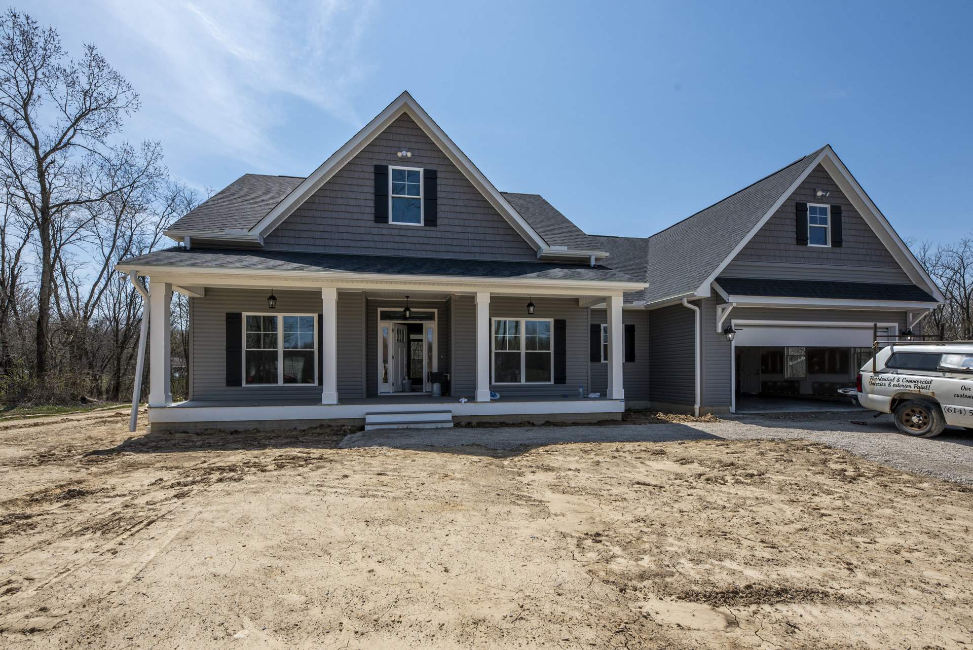 Modern white house with large windows, dirt yard in foreground, leafless tree beside home, white car with black lettering parked near entrance, reflected trees visible in window