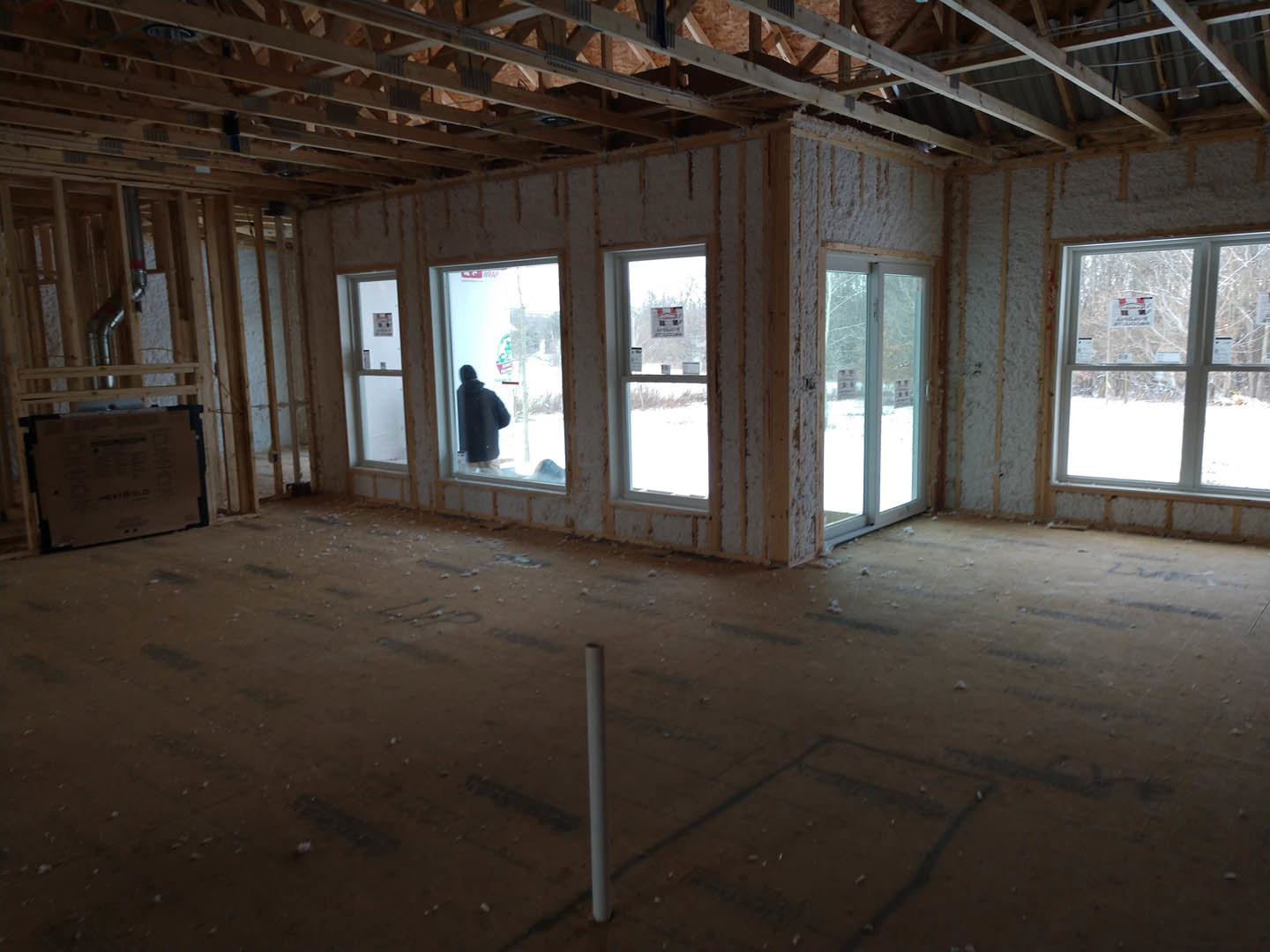 Spacious room with large windows, exposed ceiling beams, unfinished floors, white pipe, and a person in a black coat standing near a window with a red and yellow sign.