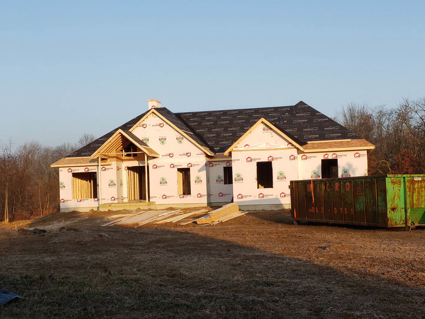 Framed house under construction with black roof and chimney, green dumpster in front, pile of lumber on dirt yard, trees and blue sky in background