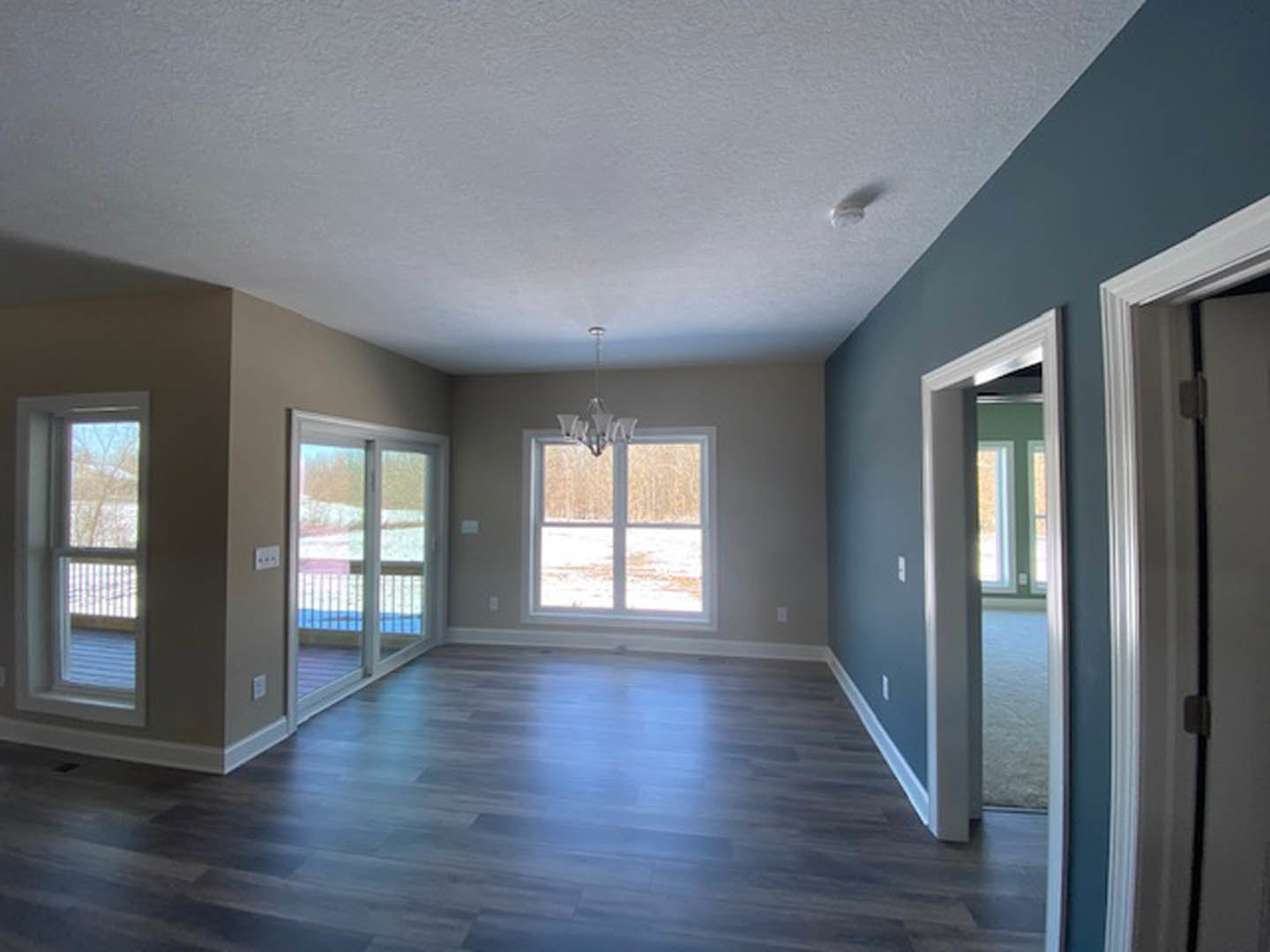Chandelier hanging from white ceiling above wide-plank wood flooring, large window with snowy landscape view, glass door with metal railing, neutral walls, doorway visible in