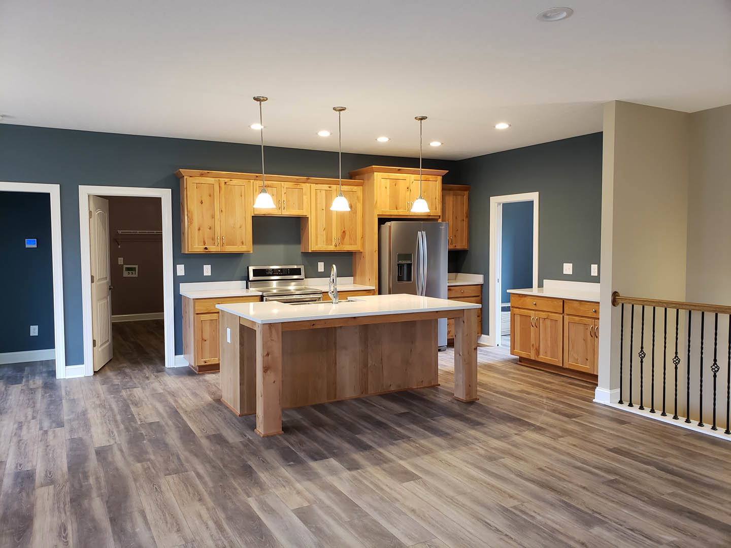 Kitchen with natural wood cabinets, spacious central island featuring a built-in sink, stainless steel refrigerator, white shelving set into a blue accent wall, open doorway