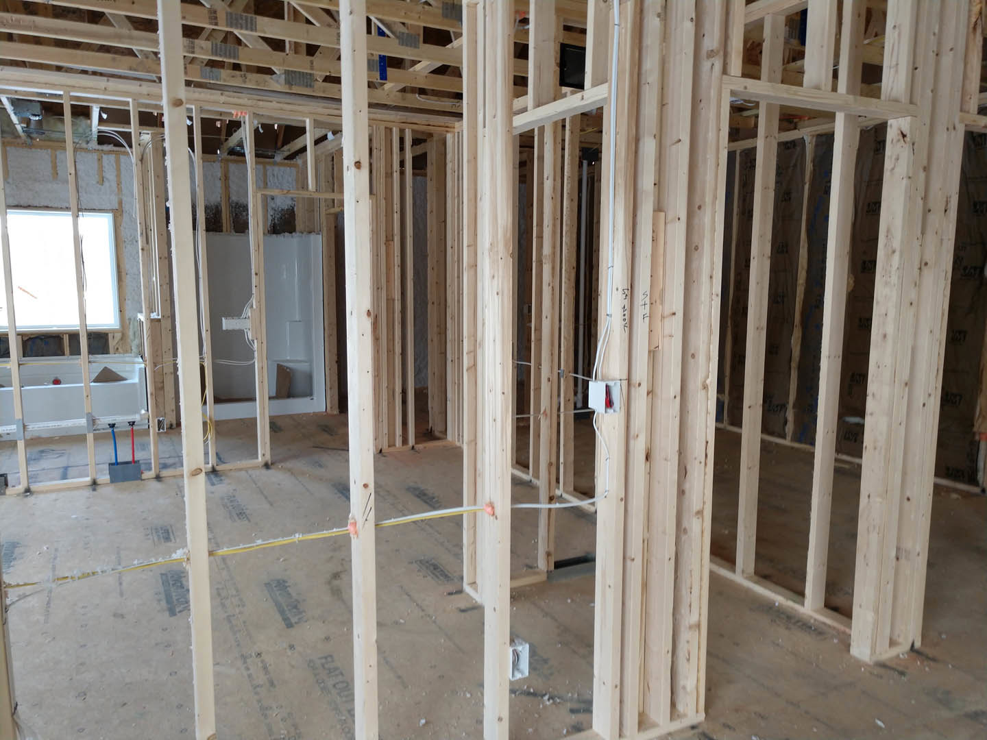 Wood framing and exposed beams in a bathroom under construction, window installed, white electrical box mounted on unfinished wall, insulation visible between lumber planks