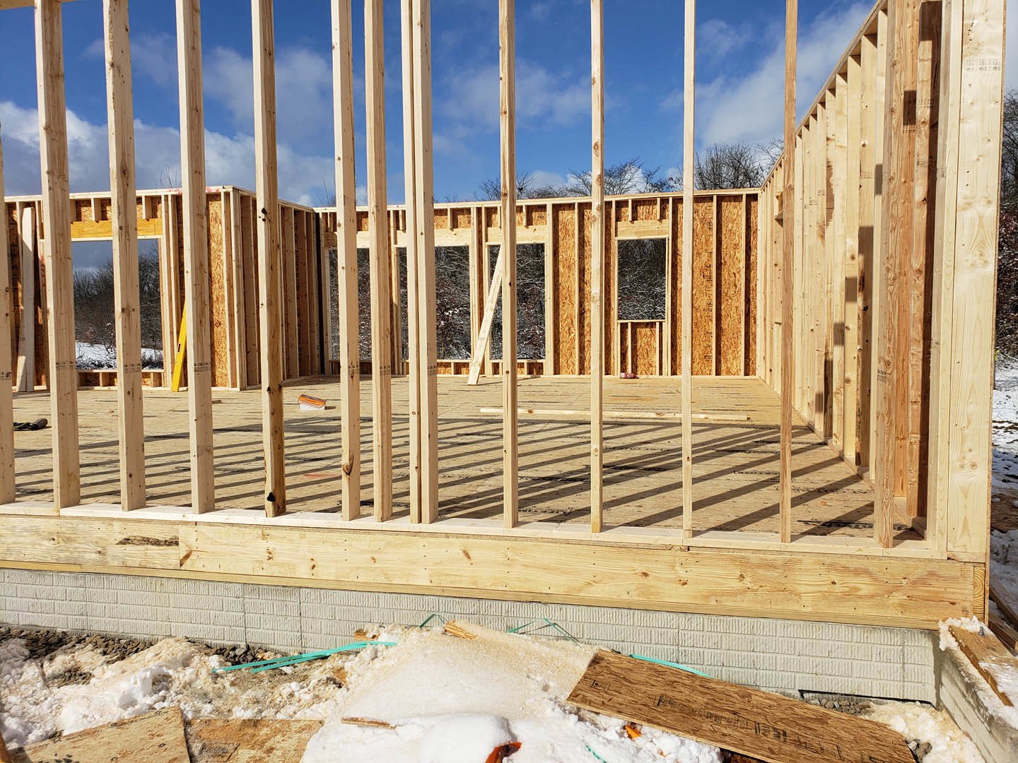 Wood-framed house under construction with exposed beams, snow covering the ground, brick wall, and partially installed window
