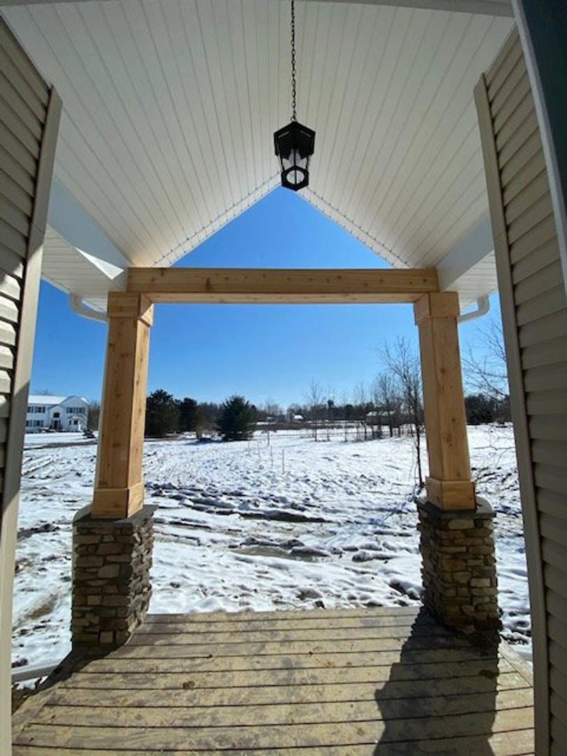 Covered front porch with ceiling light, wooden decking, stone pillars, and snow on the ground; white house exterior under clear blue sky.