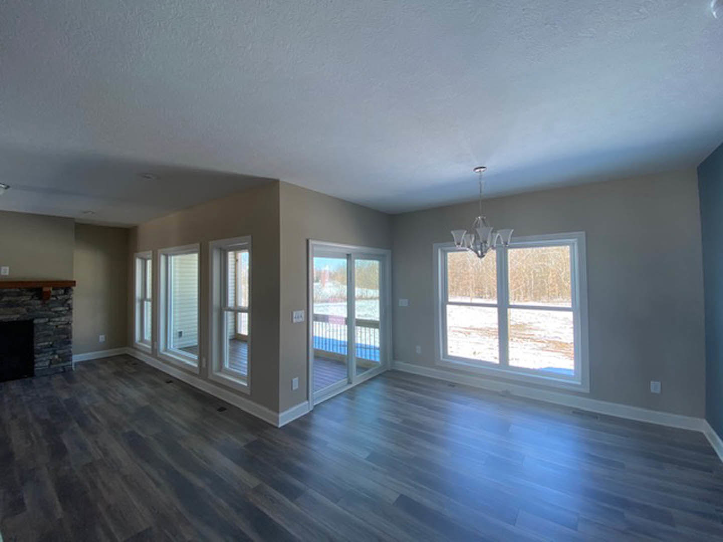 Spacious living room with large windows overlooking snowy landscape, hardwood floors, stone fireplace, modern chandelier, and sliding glass door