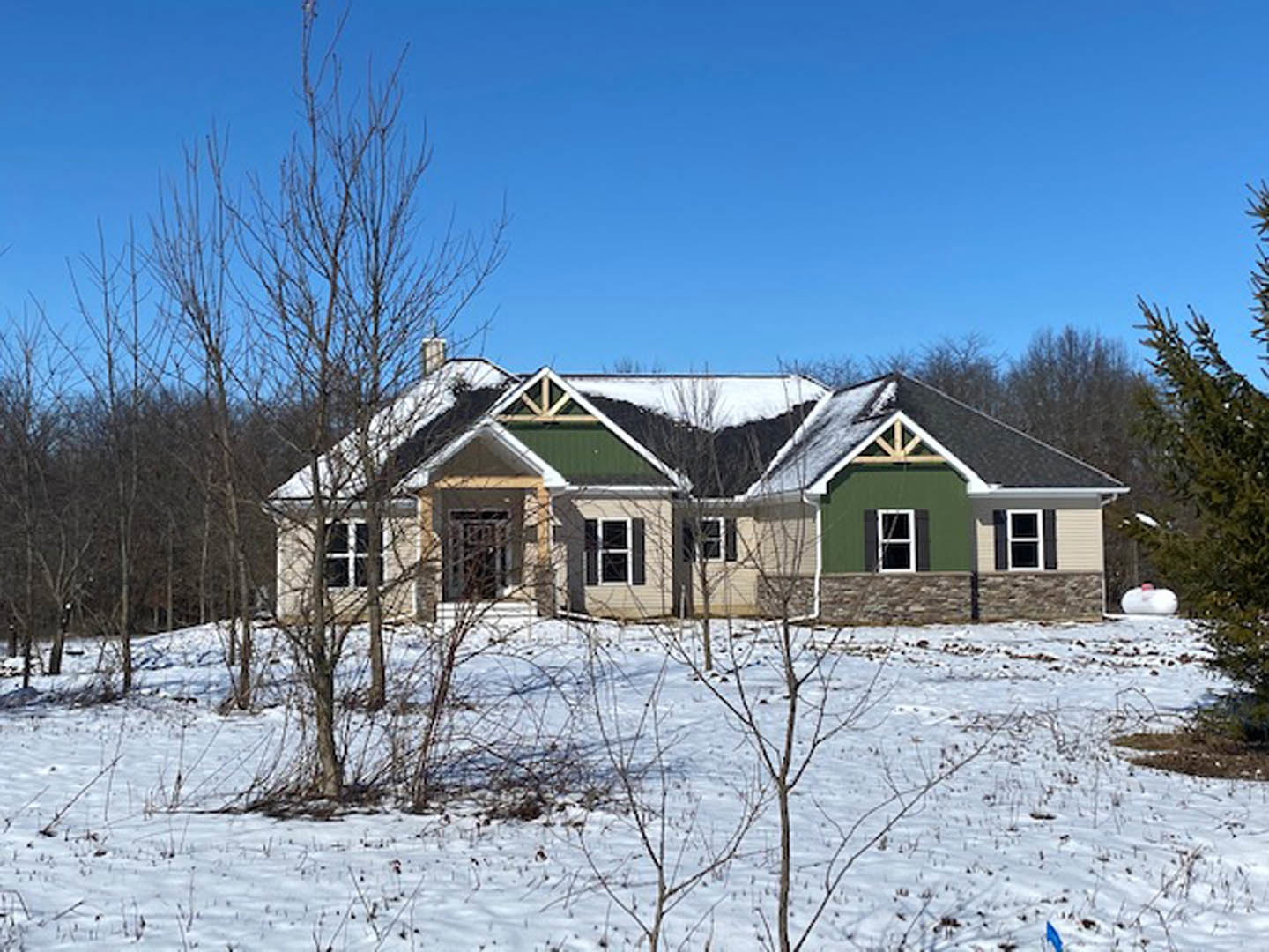 Two-story house with white-framed windows, stone wall, snow covering roof and ground, leafless trees, blue sky with scattered clouds