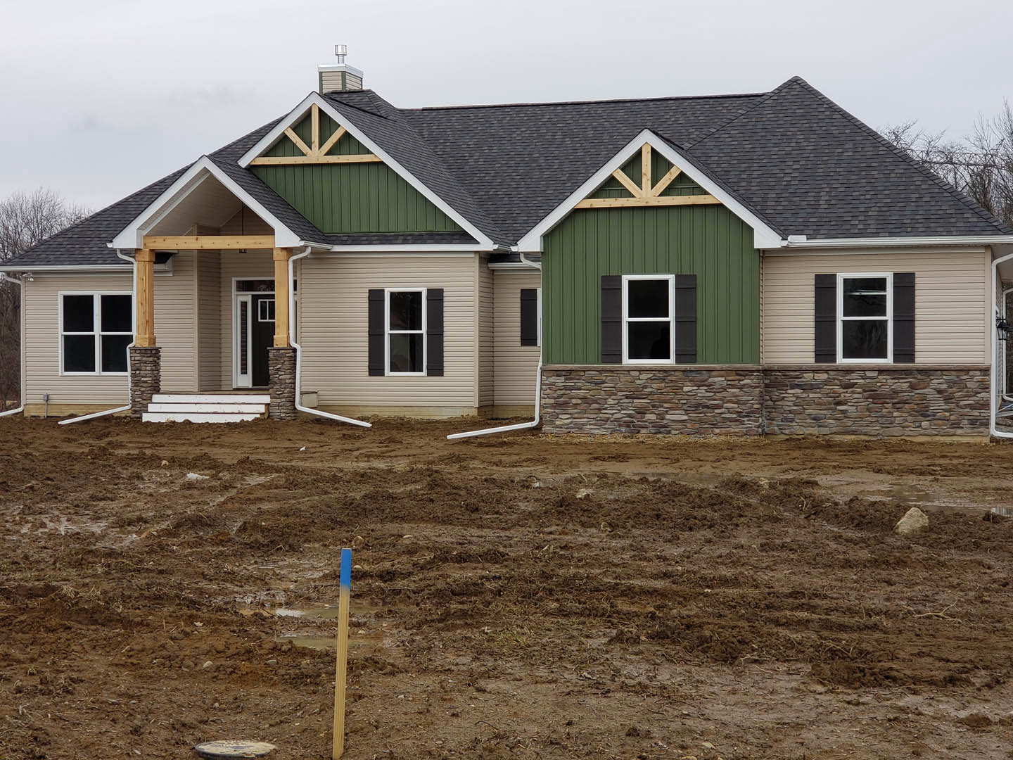 Green siding house under construction with dirt field, white framed windows with black shutters, blue and white marker in foreground, close-up of white step