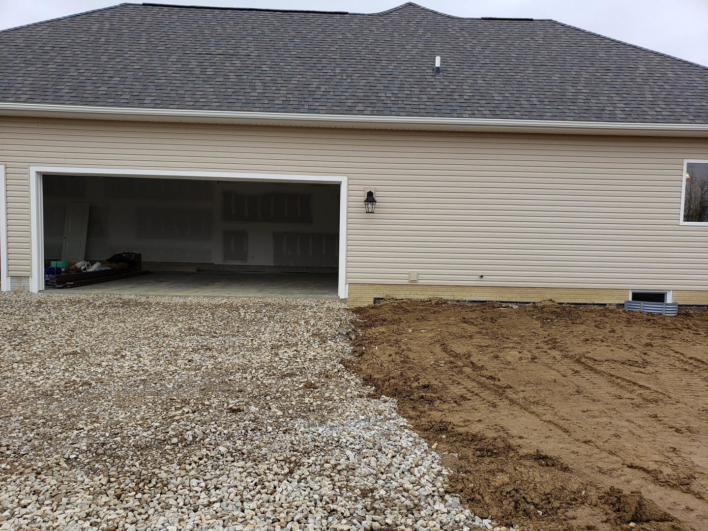 Gravel driveway leading to a garage with closed white door, lighted window, and gray siding; man seated inside garage, roof detail visible above.