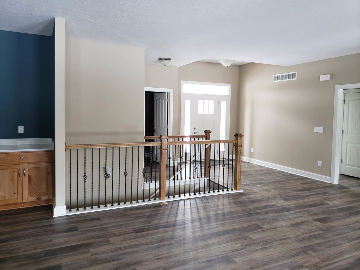 Open room featuring warm wood flooring, white door with silver doorknob, wooden railing, and built-in wooden cabinet; neutral walls and soft blue accents in the background.