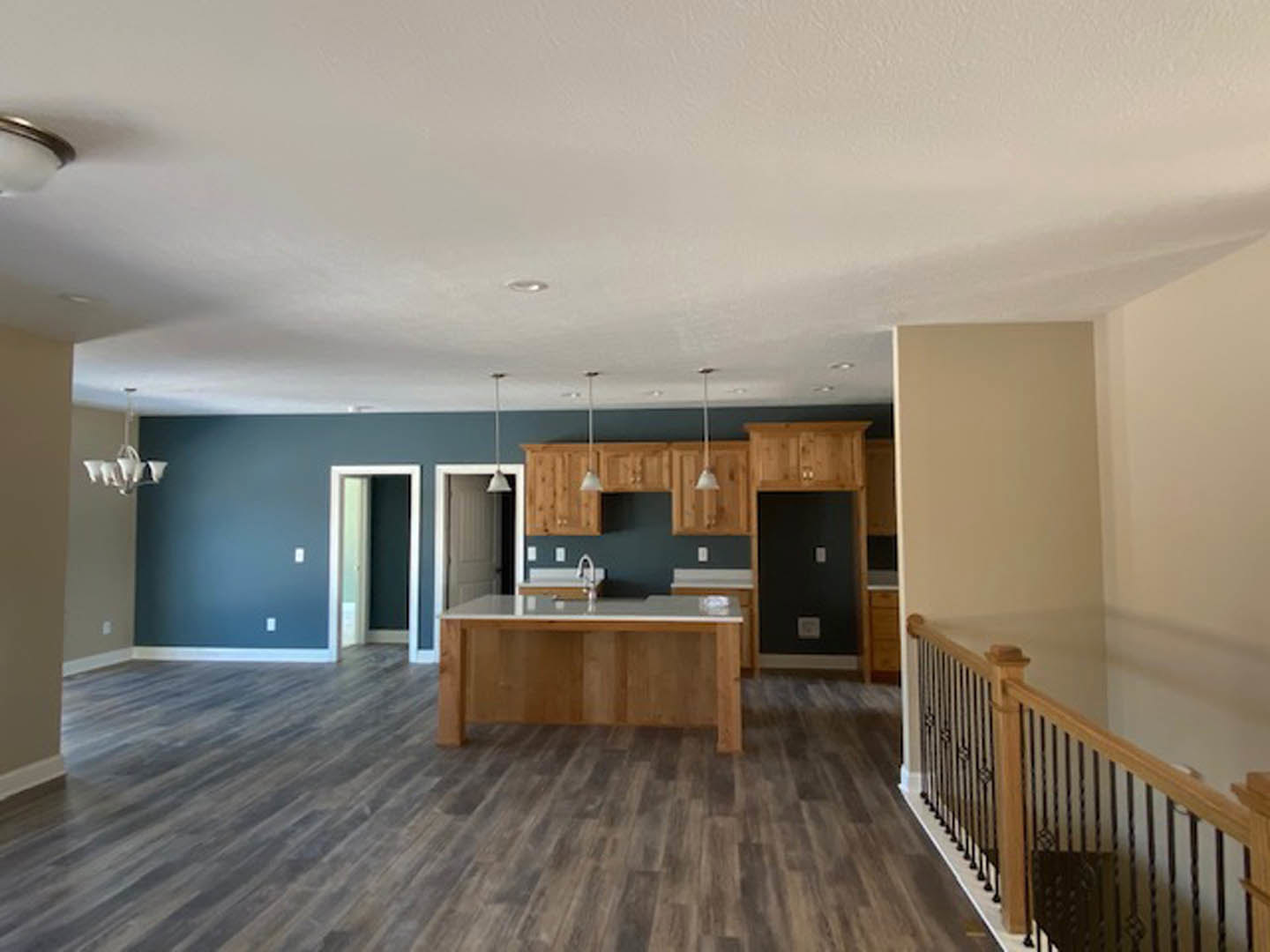 Open-concept kitchen and dining area with hardwood floors, wood dining table, white cabinetry, and modern chandelier; wooden railing and door visible in background.