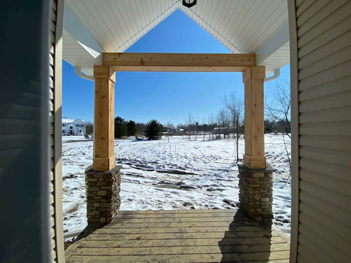 Porch with stacked stone pillars, wooden decking, and snow-covered yard; white support pole and winter landscape visible.