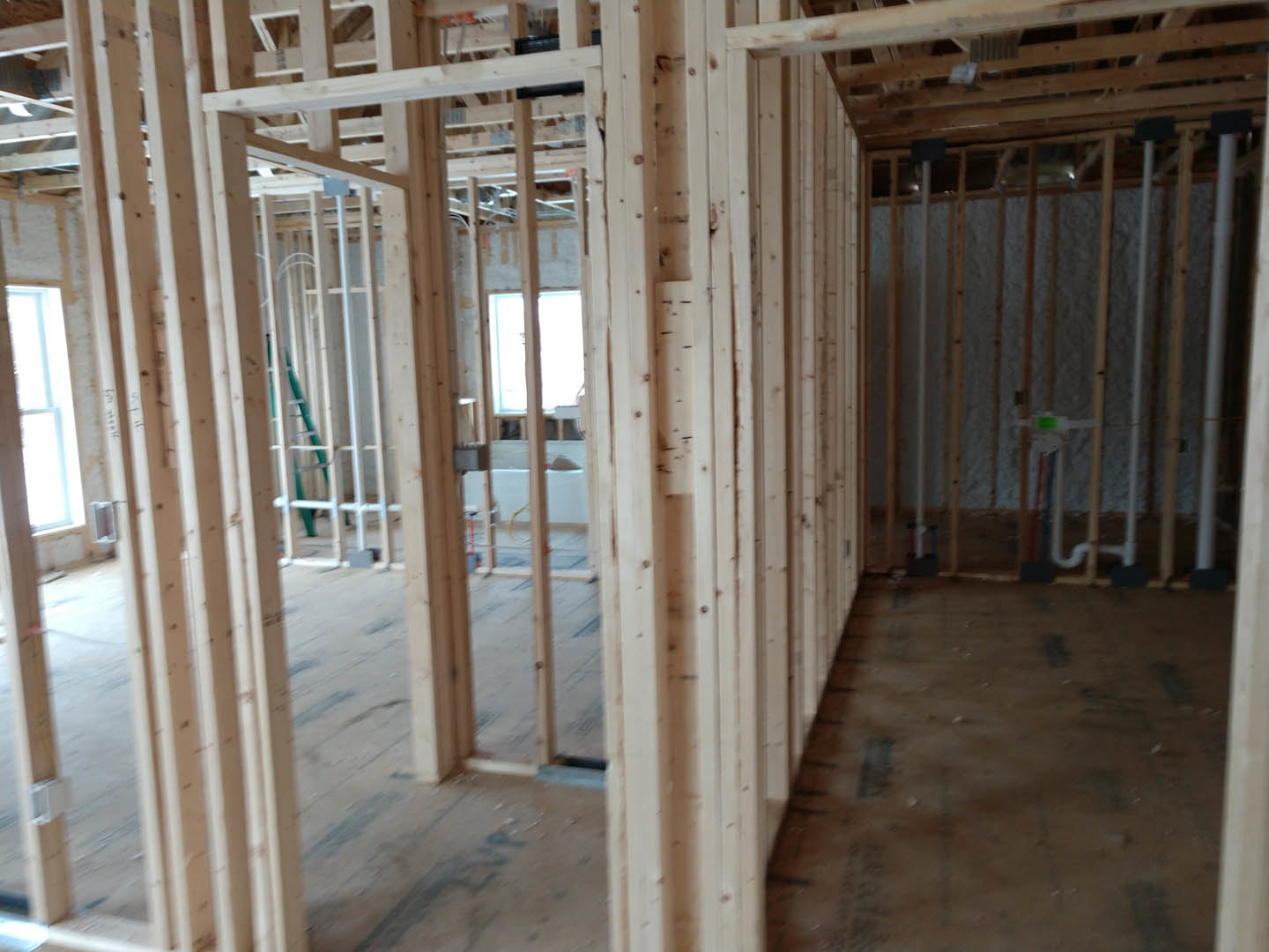 Exposed wood framing and beams in a partially constructed room, wall with visible pipes, unfinished floor, blurred ladder and flag in background