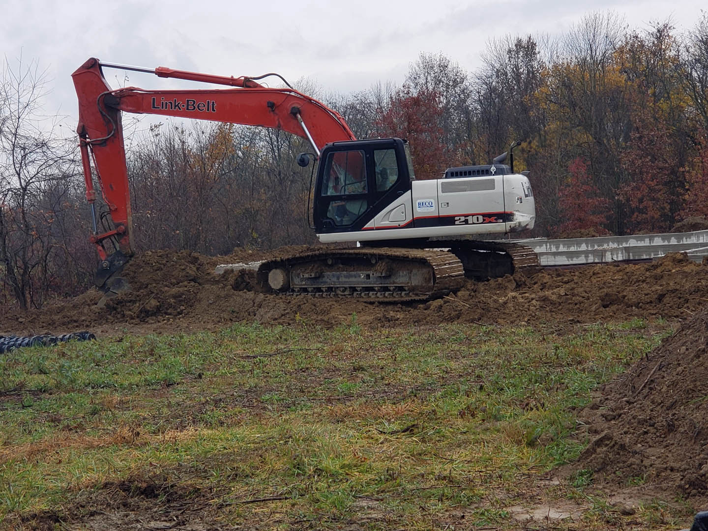 Red and white excavator with bucket digging into muddy soil on grassy construction site, surrounded by rocks and trees under open sky
