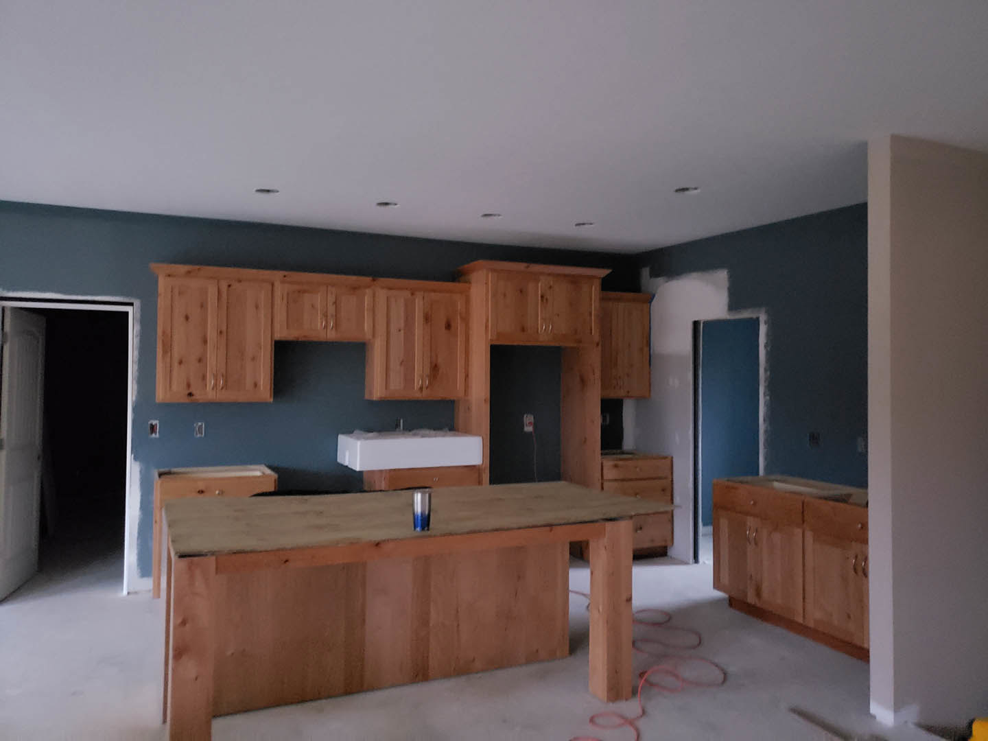 Kitchen with natural wood cabinets, stainless steel sink, white countertop, blue accent wall with white trim, cup on counter, snow visible on table outside, black door against