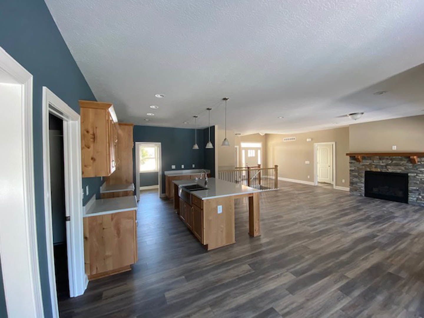 Spacious open concept living area featuring a central kitchen island with sink, brick fireplace accented by a wood beam mantel, laminate flooring, white cabinetry, and large