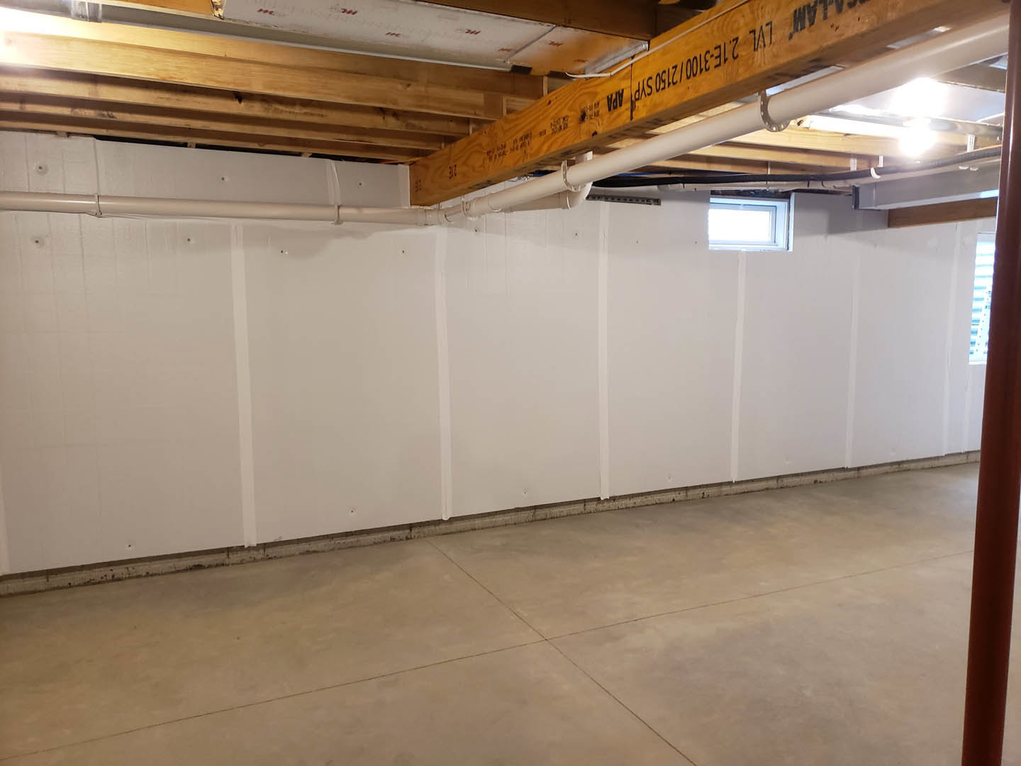 Basement with white plaster walls, wooden ceiling beams, tile flooring, and a window letting in natural light