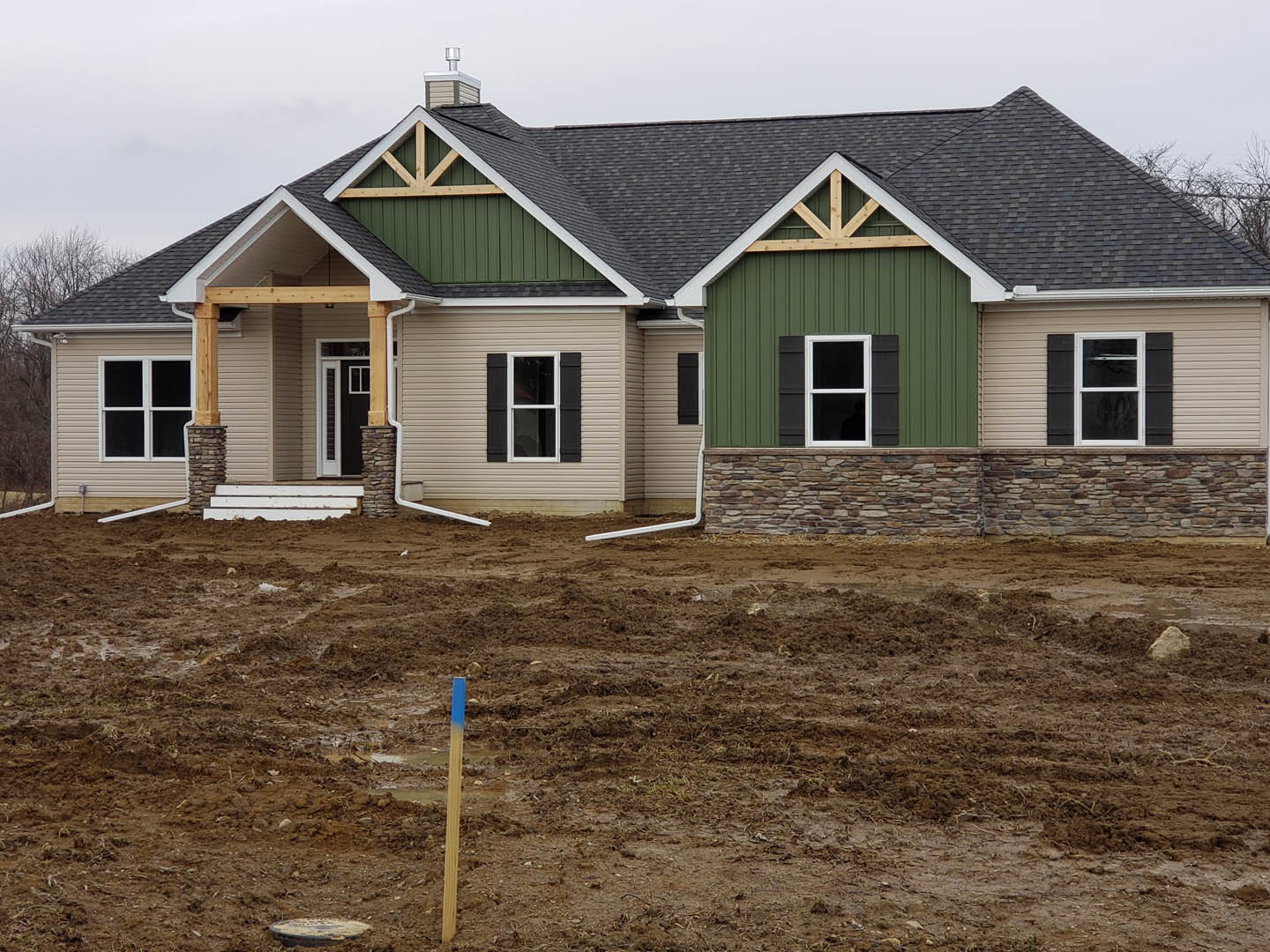 Partially built home with green roof, white-framed window, exposed siding, dirt yard featuring a blue and white pole, scattered rocks, and construction post