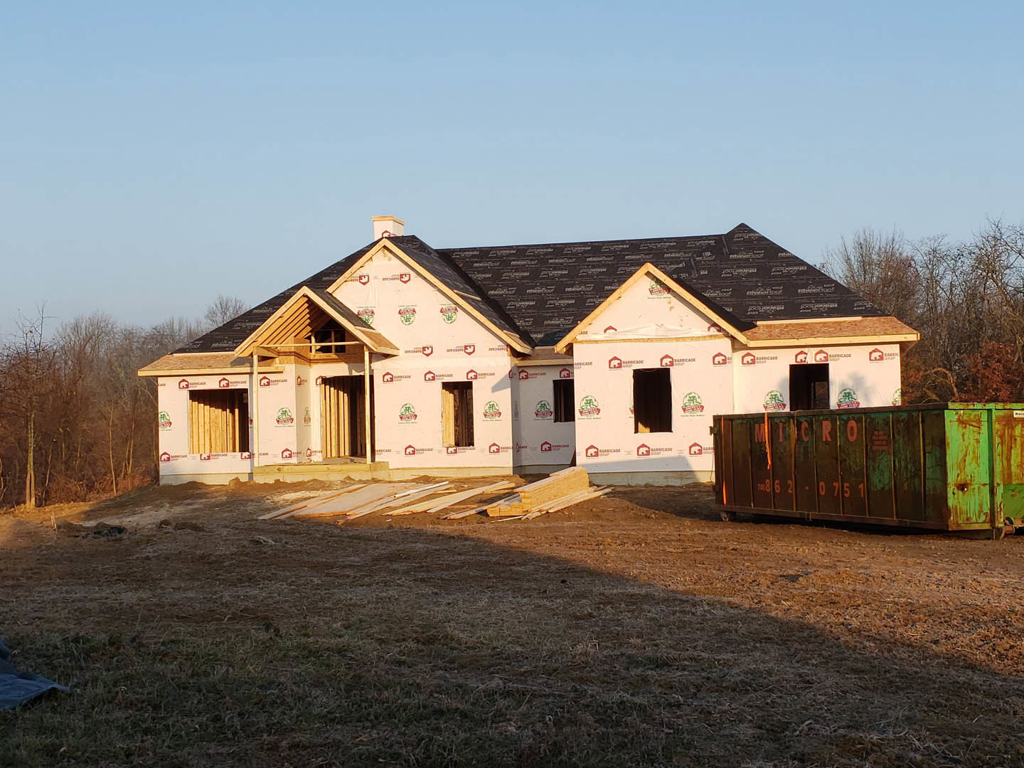 Partially built house with black shingle roof, exposed framing, green dumpster in driveway, scattered lumber in yard, trees in background