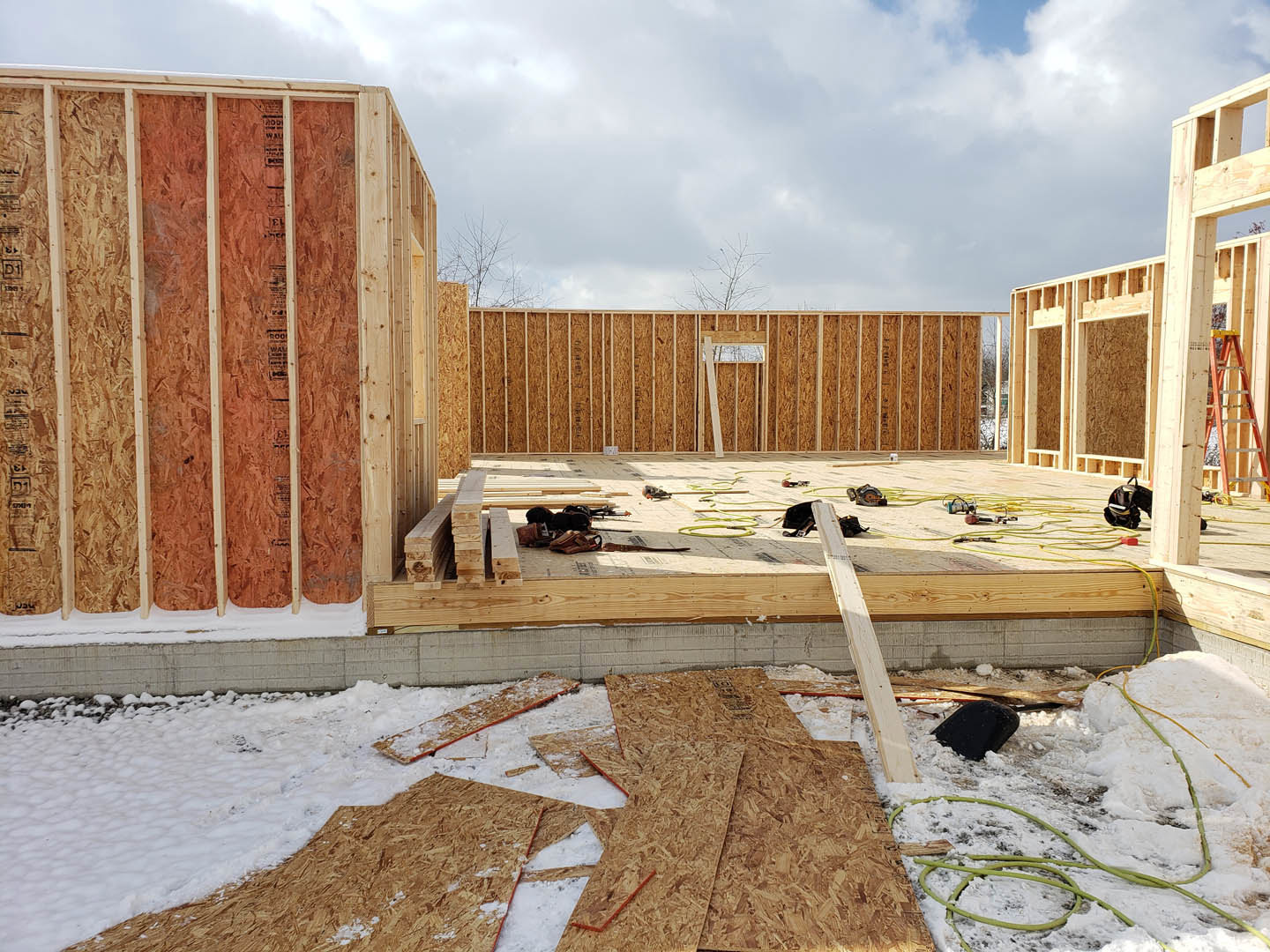 Wood-framed exterior wall under construction, snow covering ground, scattered wooden planks, black and red painted wood, cloudy sky overhead