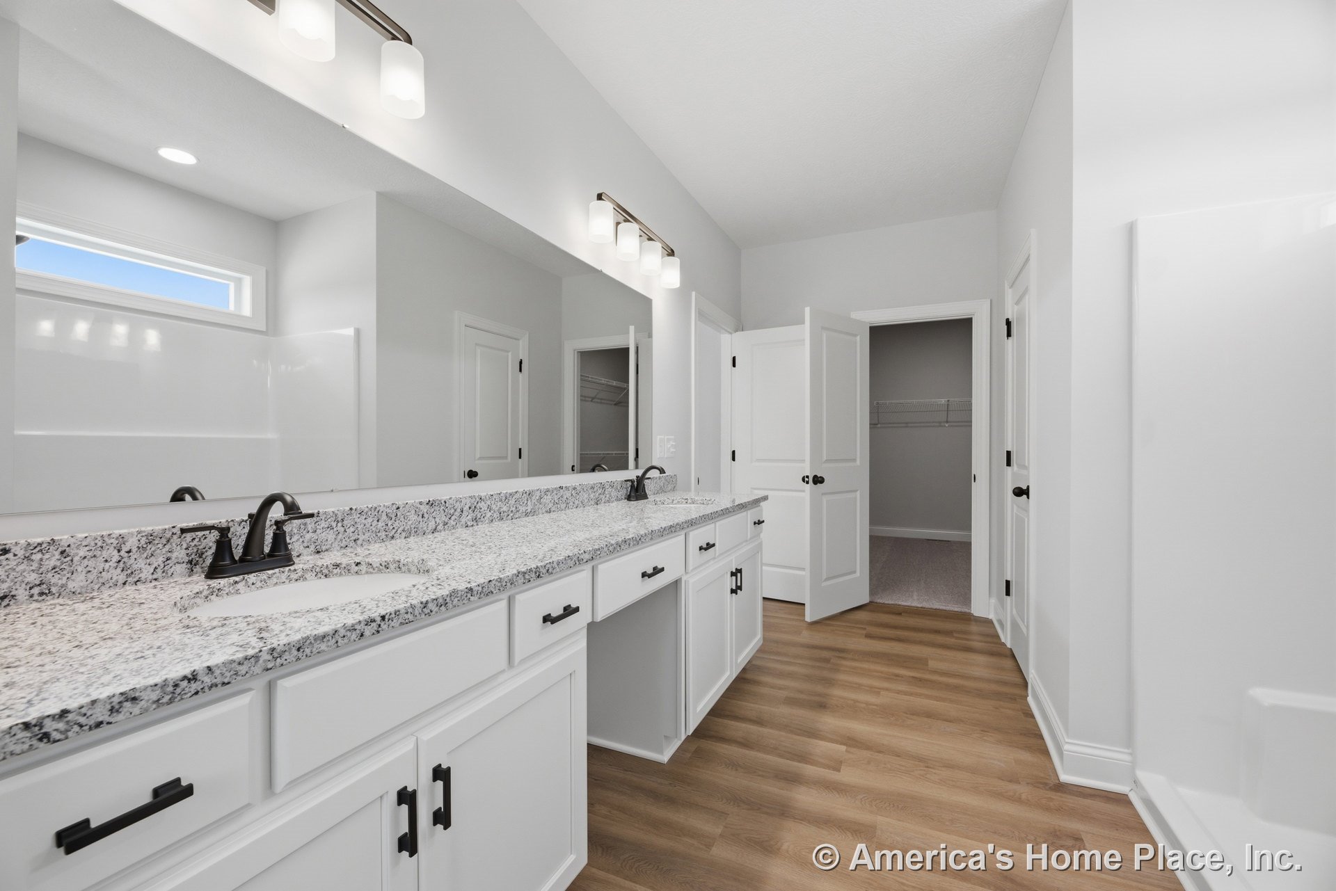 Double vanity with granite countertop, white shaker cabinets, undermount sinks, black hardware, large wall mirror, built-in overhead lighting, wood-look flooring, white trim, door