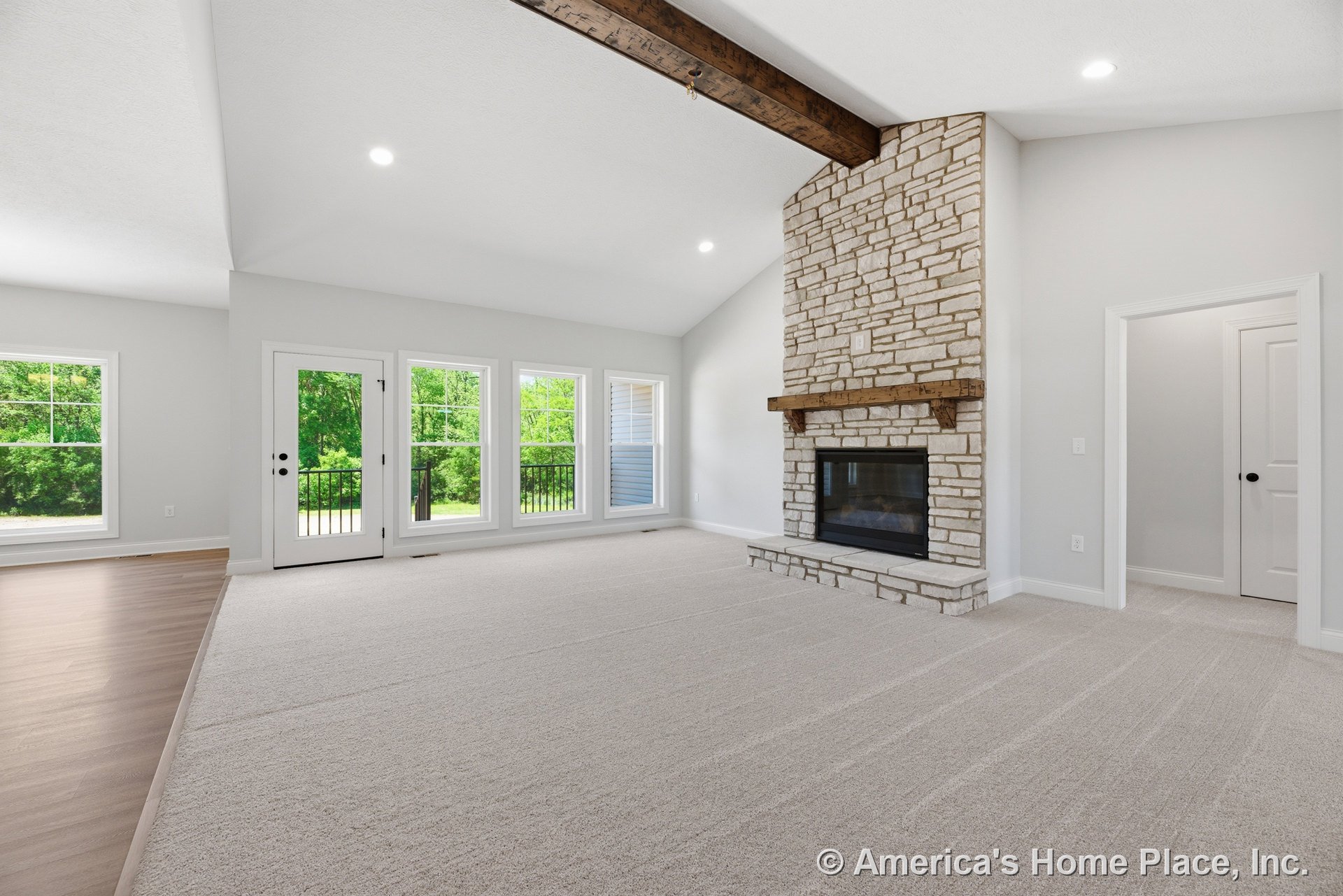 Vaulted living room ceiling with exposed wood beam, stone fireplace, large windows, carpet flooring, white trim, recessed lighting, and open layout.