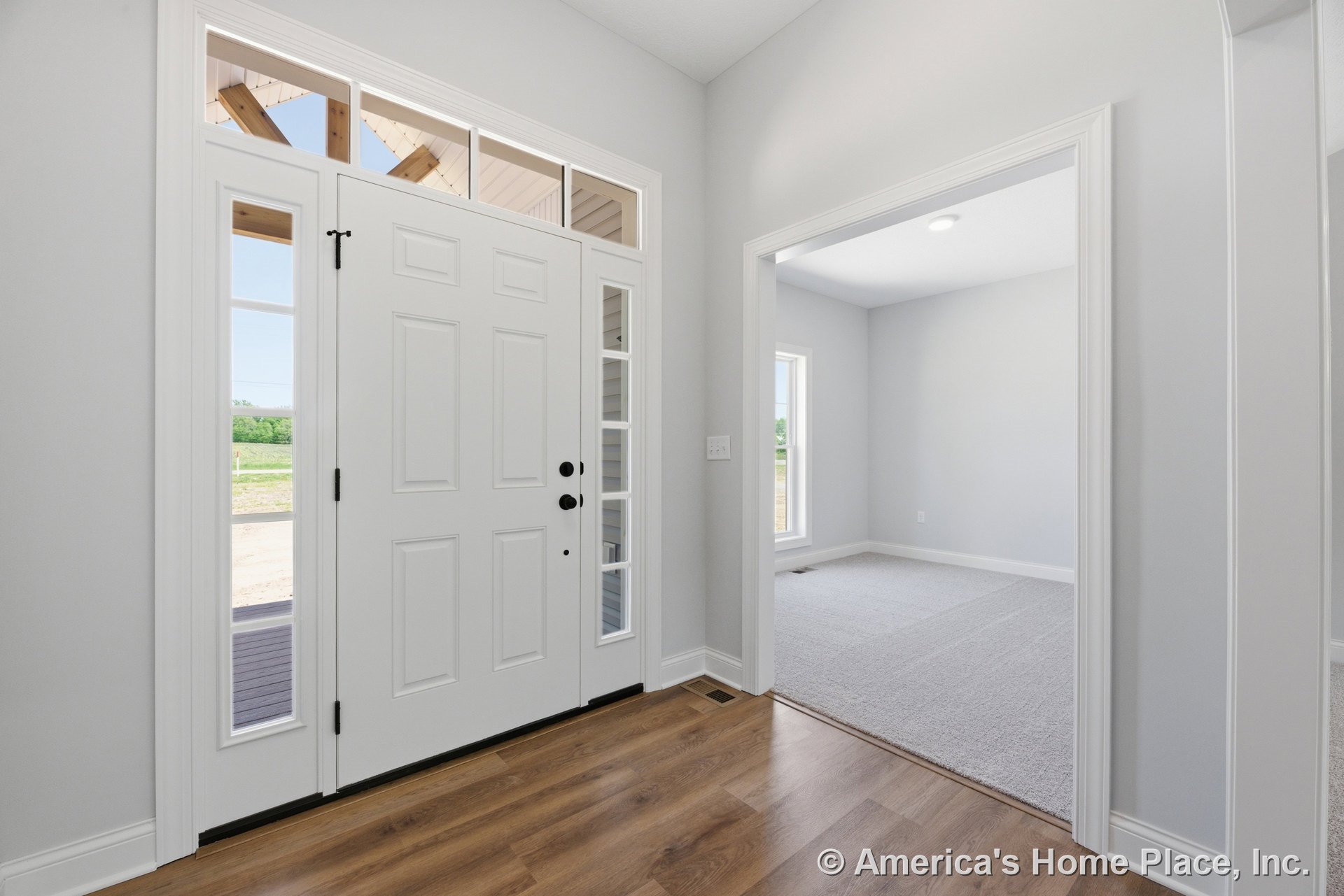 White paneled front door with sidelights and transom windows, wood-look flooring in foyer transitioning to carpeted adjacent room, large trim and baseboards, recessed ceiling