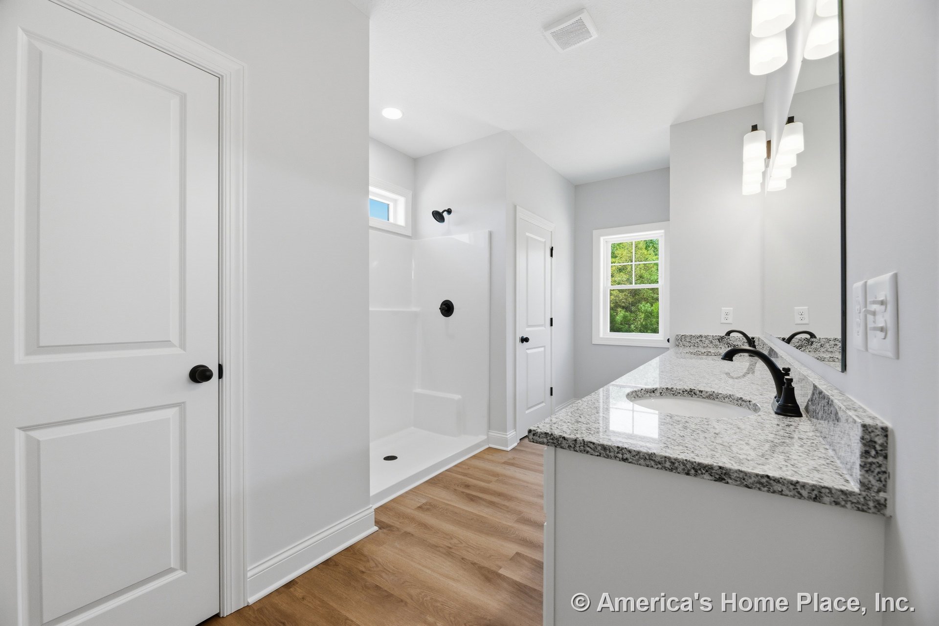 Double vanity with granite countertops and black fixtures, walk-in shower with glass enclosure, wood-look flooring, large wall mirror, modern overhead lighting, white trim and