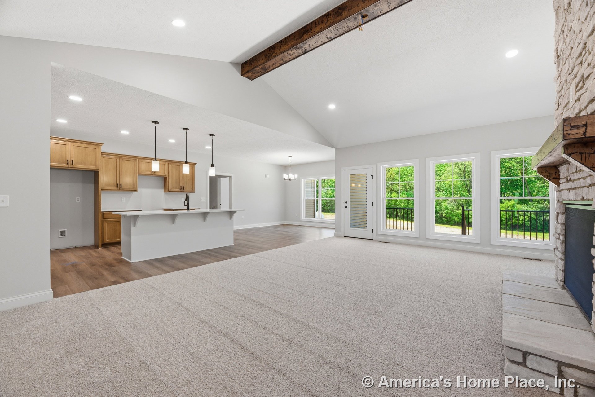 Vaulted ceiling with exposed wood beam, stone fireplace with mantel, wall of large windows with white trim, open kitchen featuring wood cabinetry and pendant lights above island