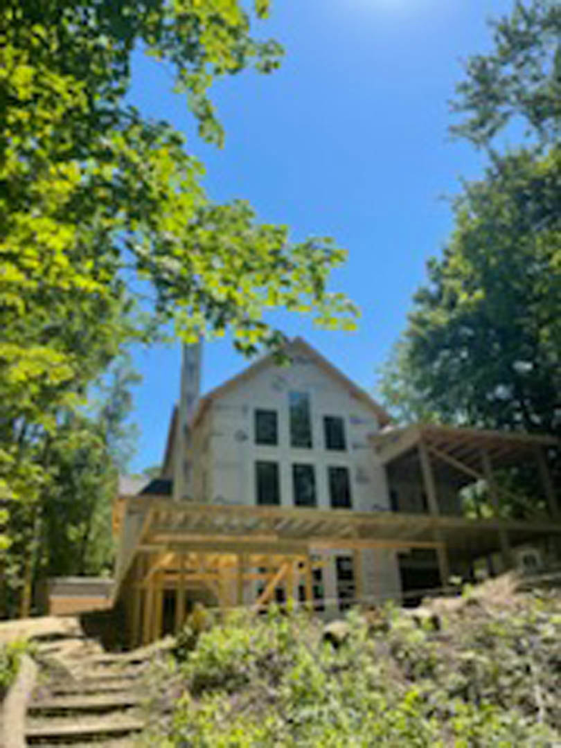 Partially built custom home with exposed framing, unfinished exterior walls, and surrounding mature trees in the background