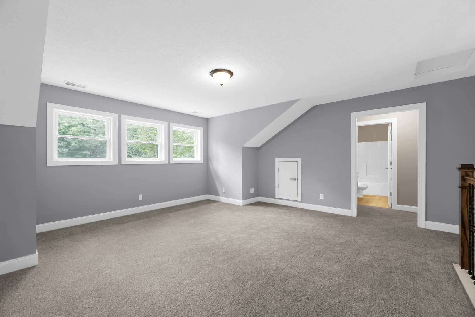 Carpeted room with white walls, large windows, white door with silver handle, ceiling light fixture, and adjacent bathroom with toilet and door