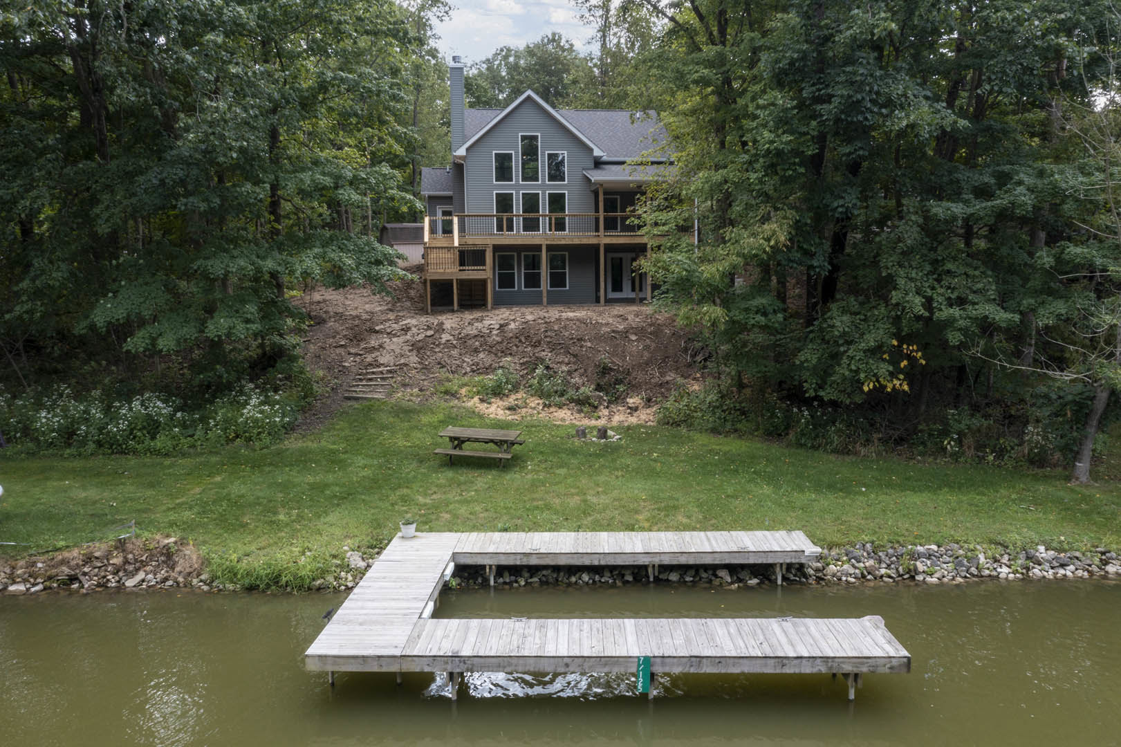 Two-story house with gray siding and white trim, surrounded by trees and grass, wooden dock extending over pond, picnic table on lawn near water