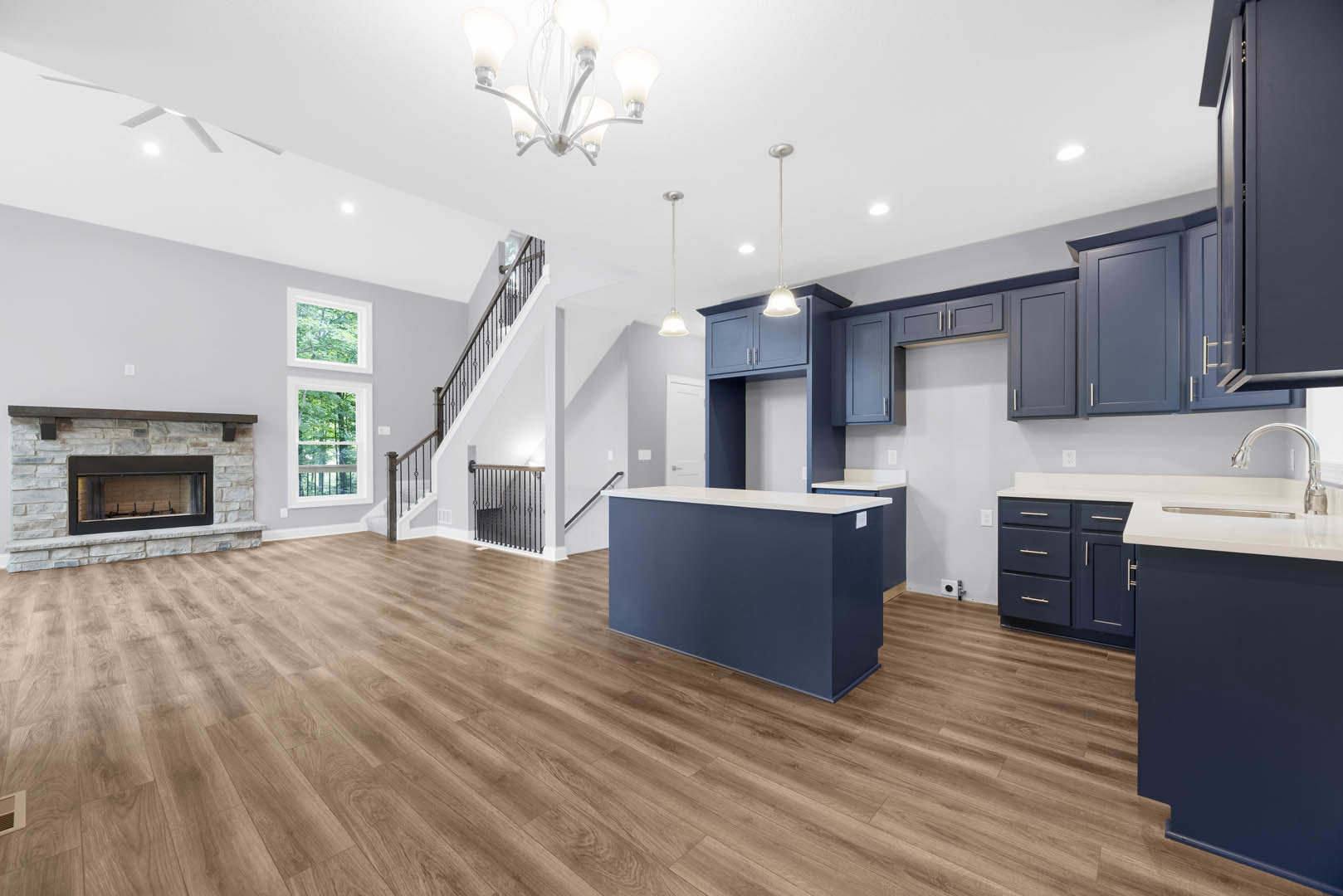 Open kitchen with blue island, white quartz countertop, hardwood flooring, modern chandelier, blue accent wall, glass-front fireplace, person standing near fireplace